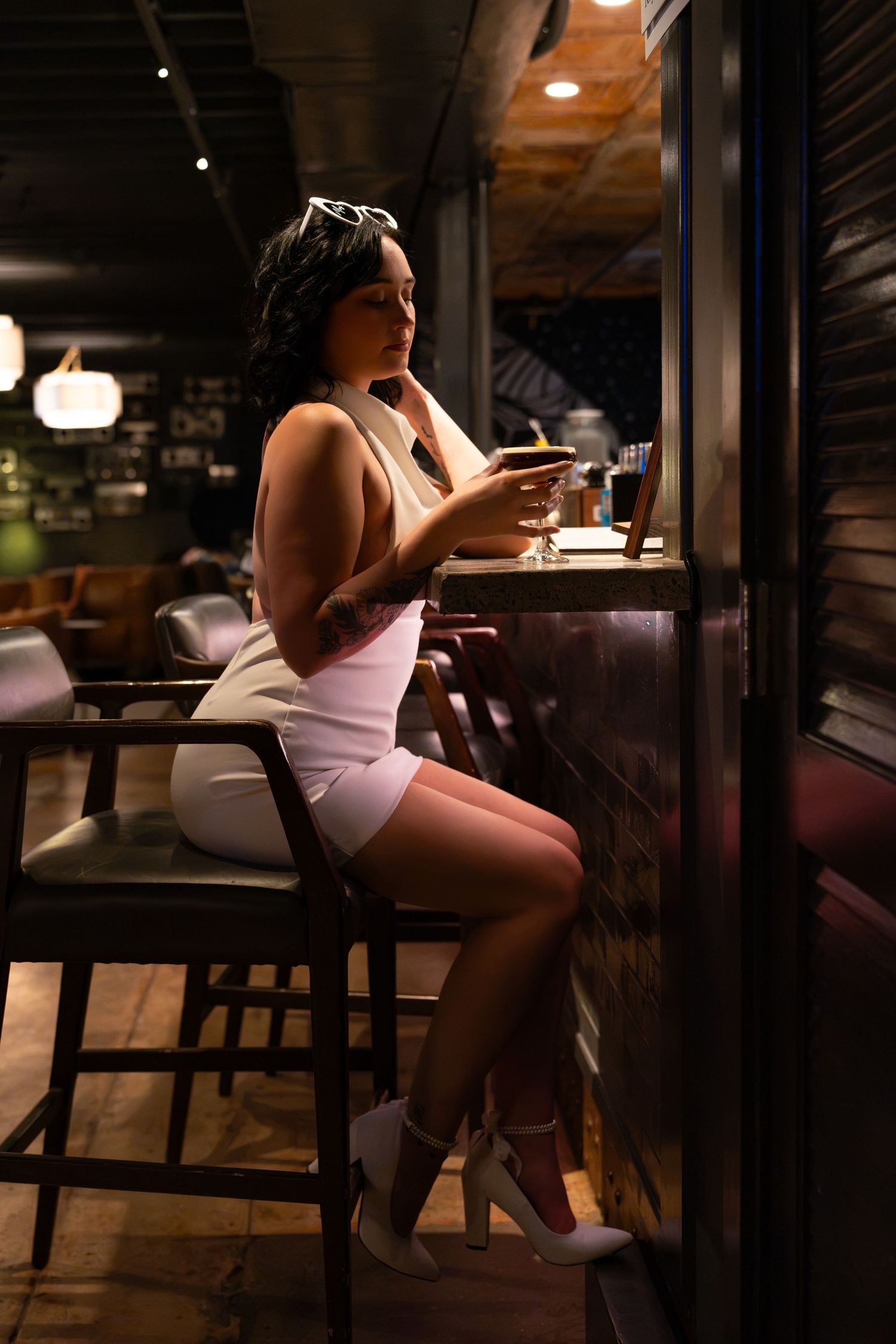 Woman in a white mini dress and heels sits on a leather barstool sipping a drink at a dimly lit bar with warm ambient lighting.