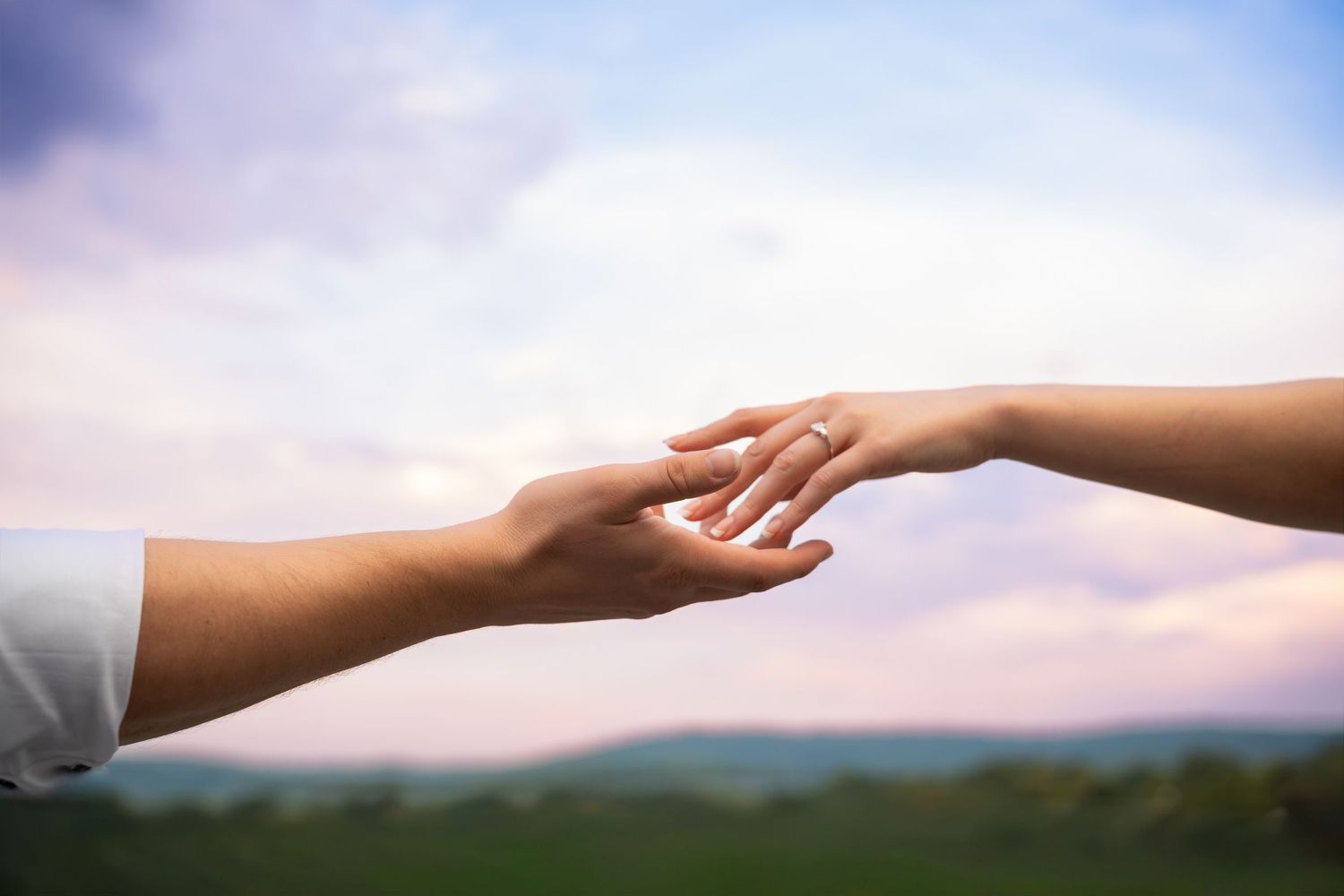 Two hands reaching toward each other against a soft pastel sky and green rolling hills, symbolizing connection, support and compassion.