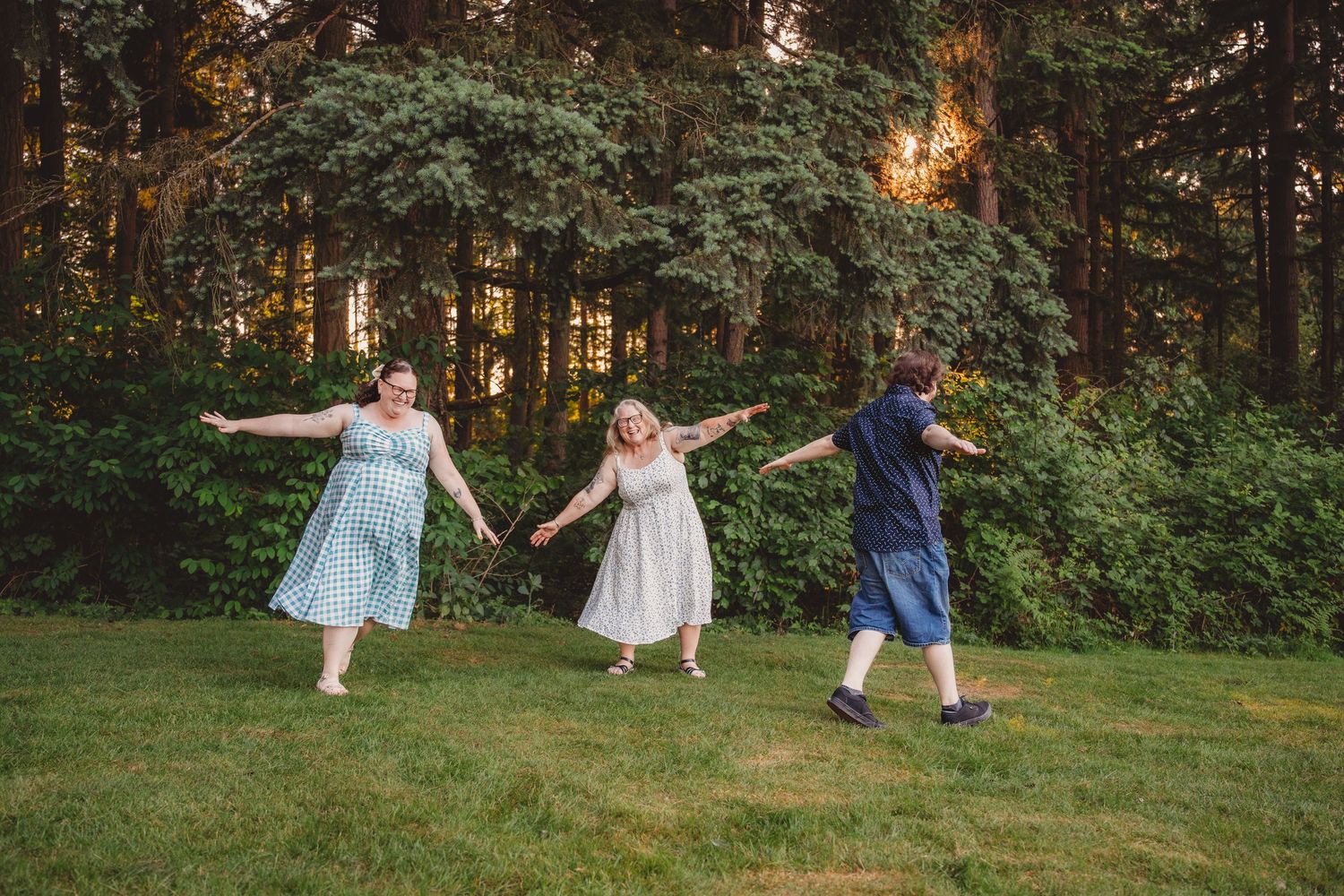 Three women in dresses hold hands and dance together on a green lawn surrounded by tall trees at sunset.