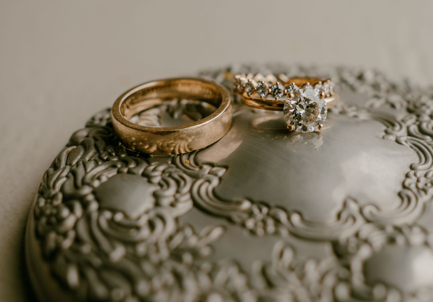 Two gold wedding rings, one plain band and one diamond crown ring, resting on an ornate silver trinket dish.