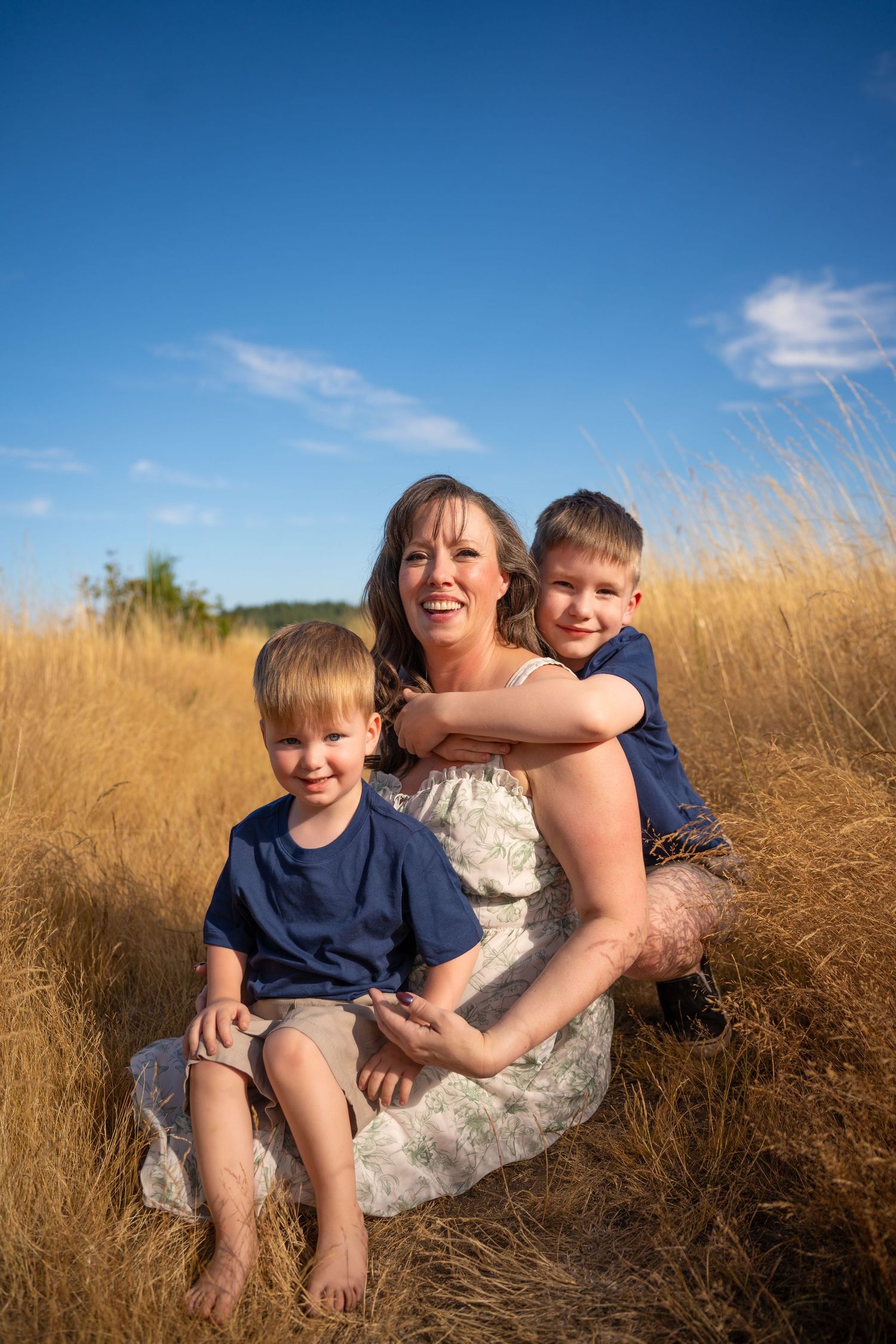 A mother sits in golden grass with her two young sons in blue shirts under a bright blue sky.