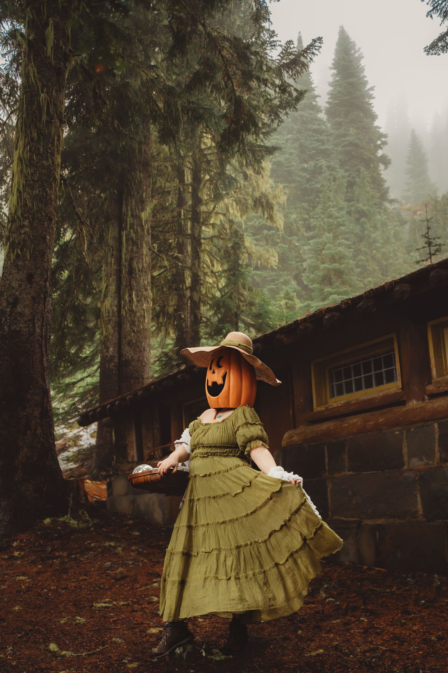 Person in green ruffled dress with jack-o-lantern head and hat poses in misty forest near rustic cabin.