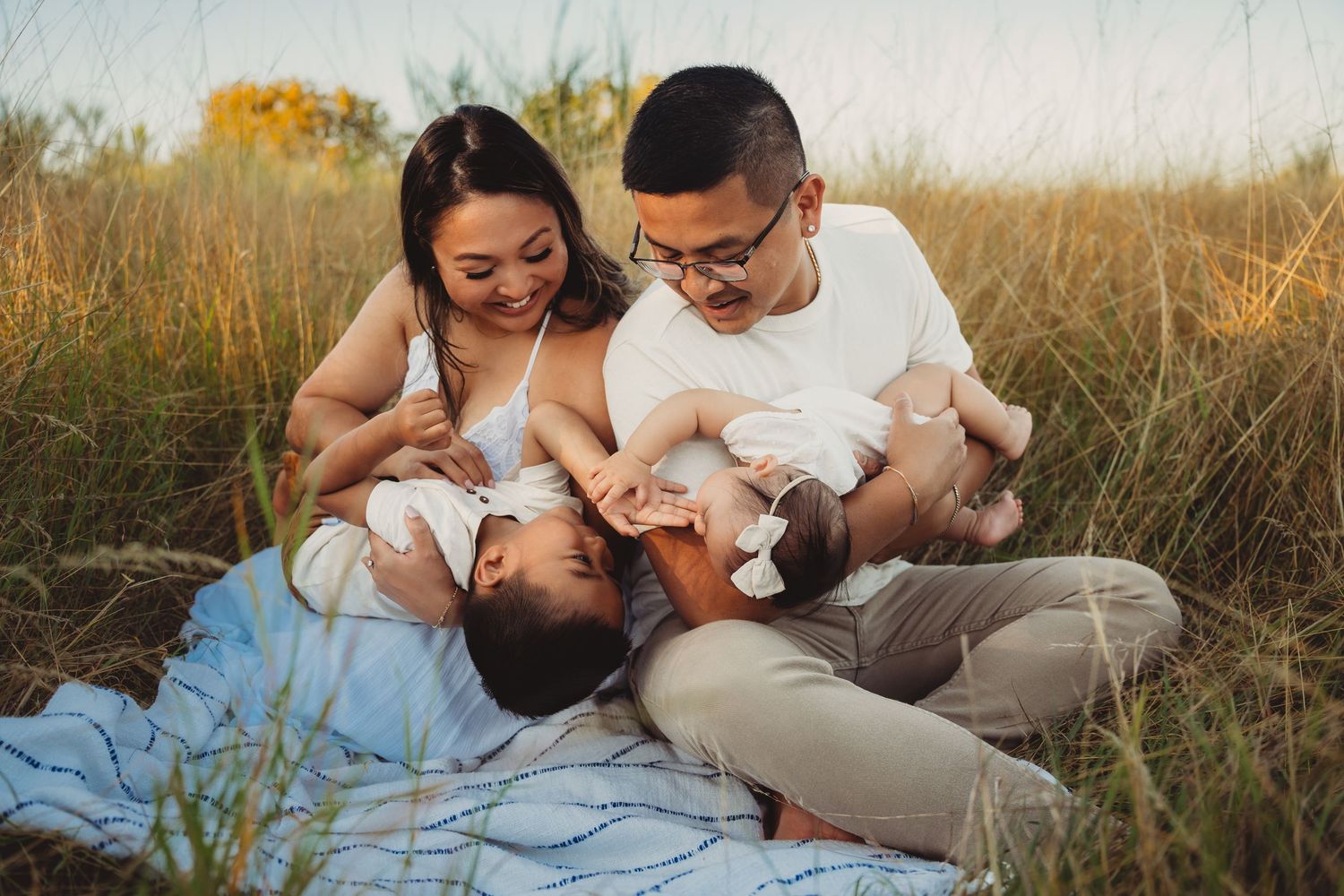 A young Asian family sits on a blanket in a golden field, parents smiling while holding two babies outdoors.