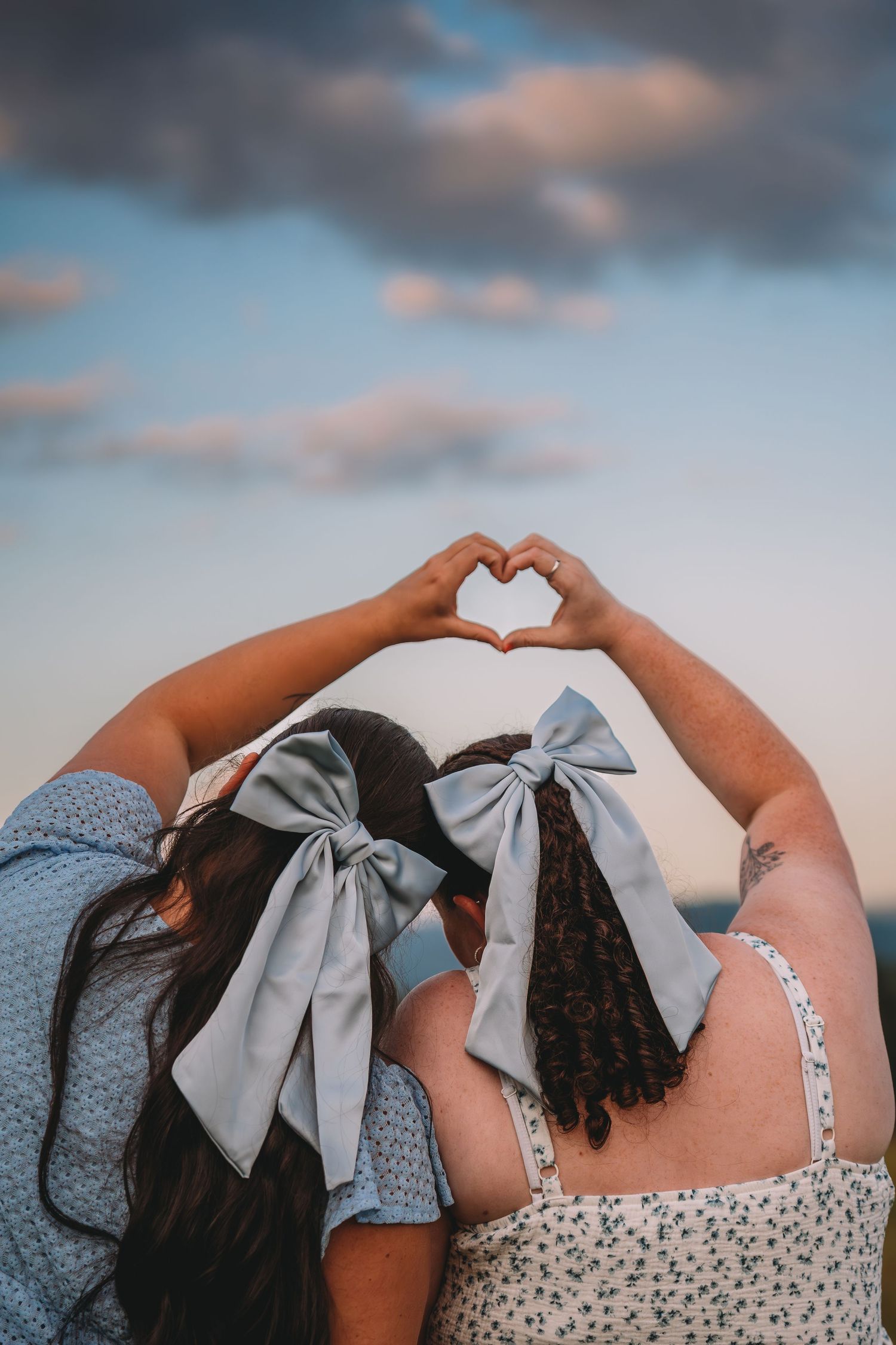Two women with gray bow hair accessories make a heart shape with their hands against a cloudy sunset sky.