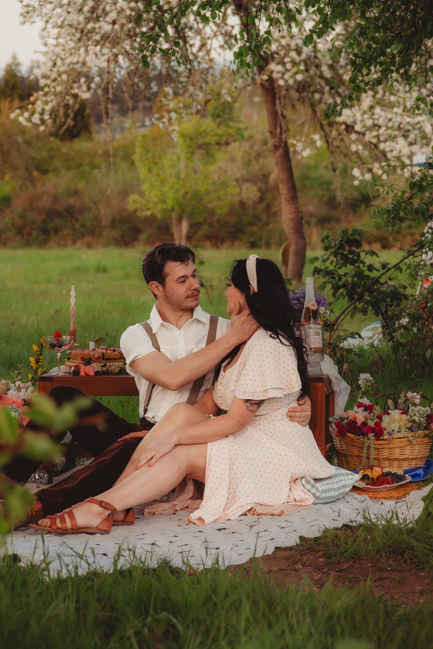 Couple sharing a romantic outdoor picnic on a blanket under blooming trees in a lush green meadow.
