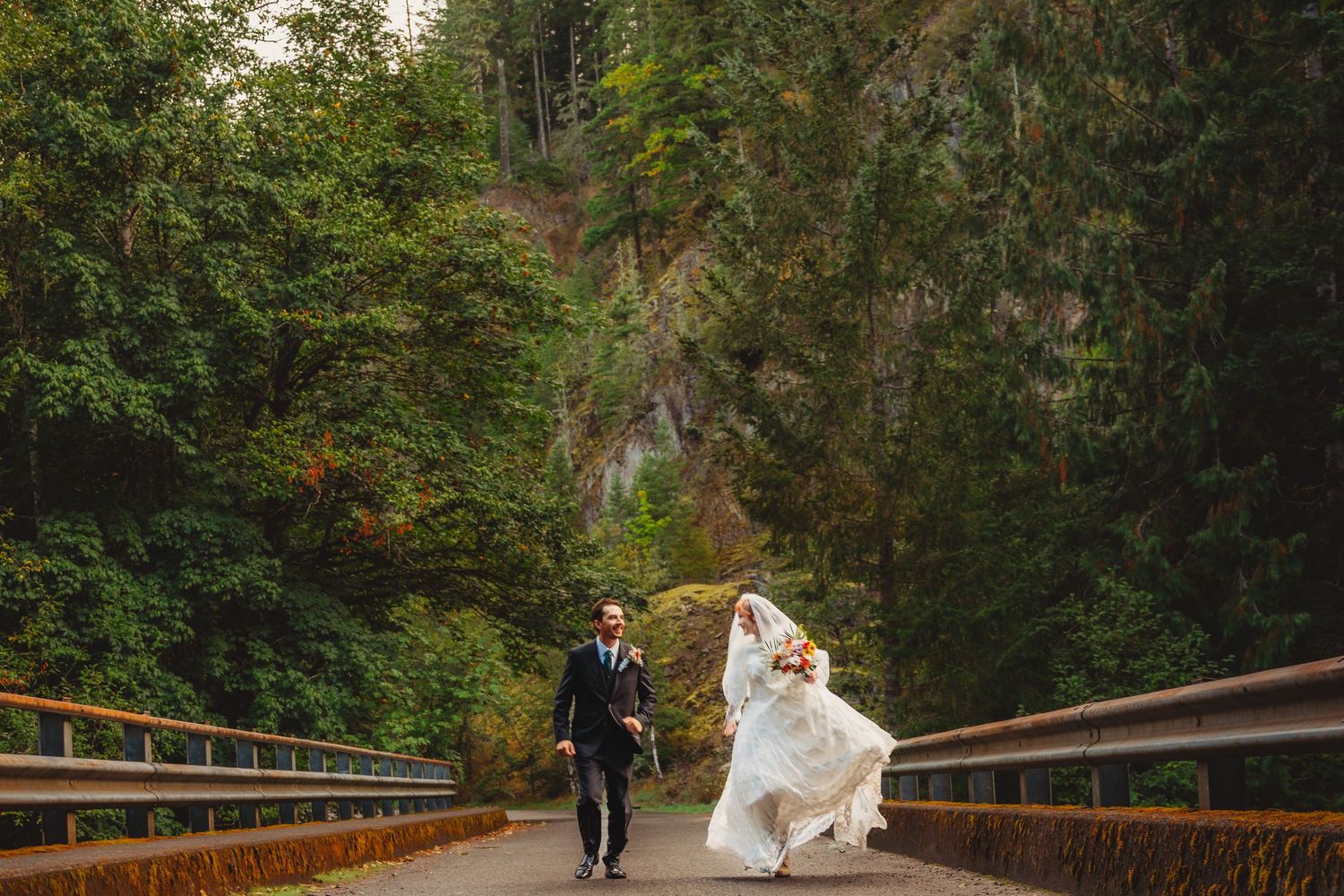 Bride and groom walking hand in hand on a bridge surrounded by lush green forest on their wedding day.
