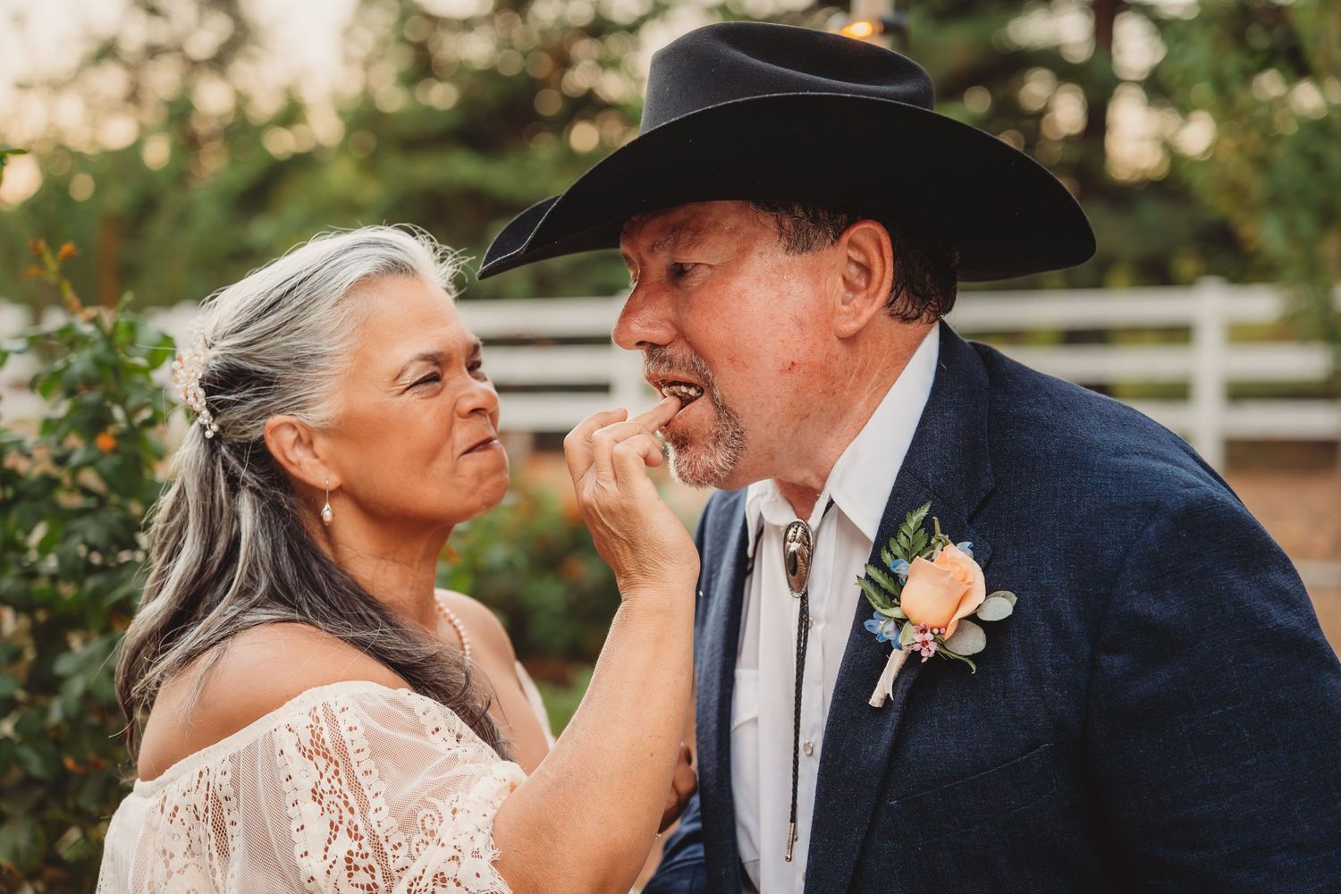 Older bride feeding cake to groom in cowboy hat at rustic outdoor wedding celebration with white fence backdrop. California