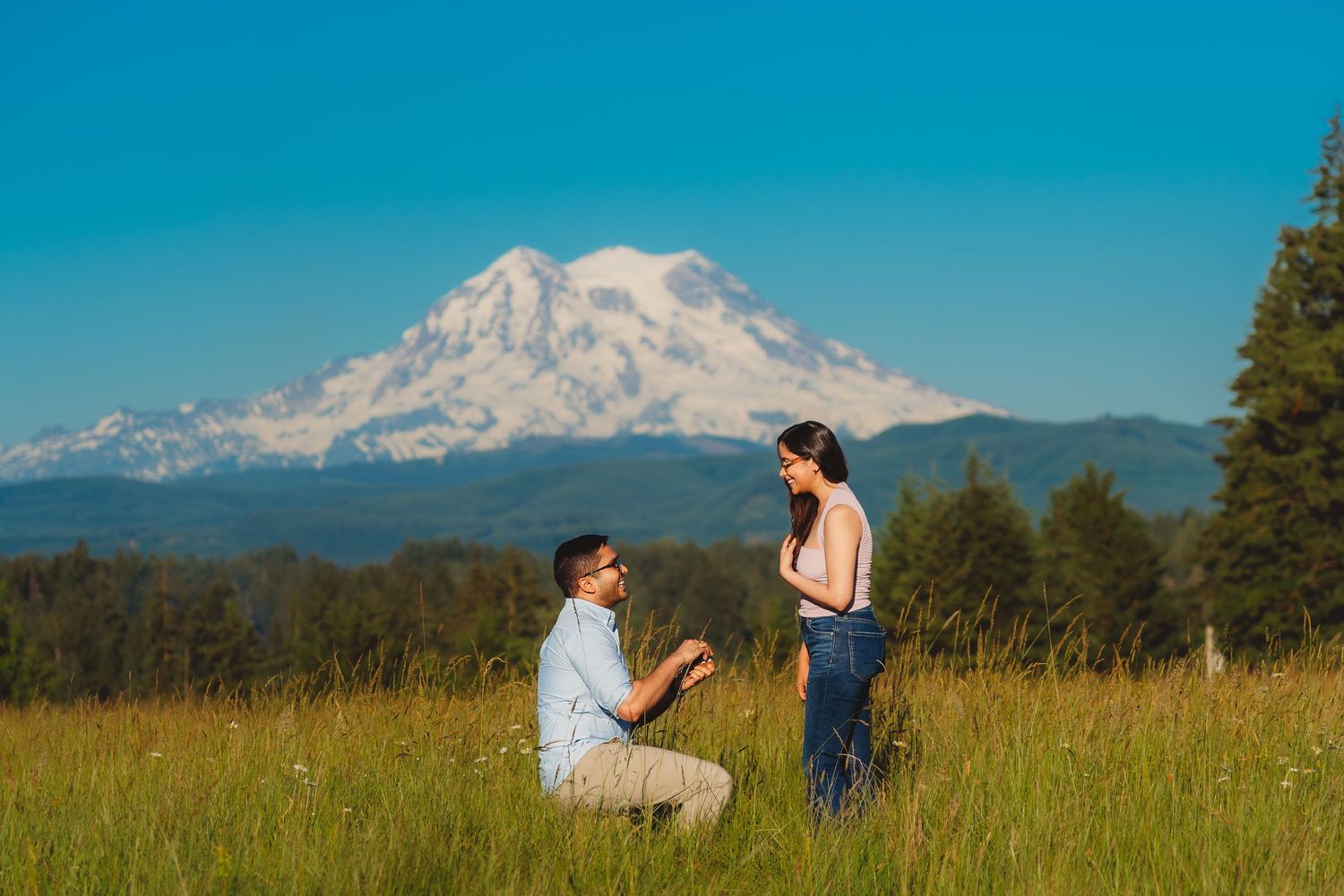 Man proposes to woman in sunny meadow with snow-capped Mount Rainier in background on clear blue sky day.