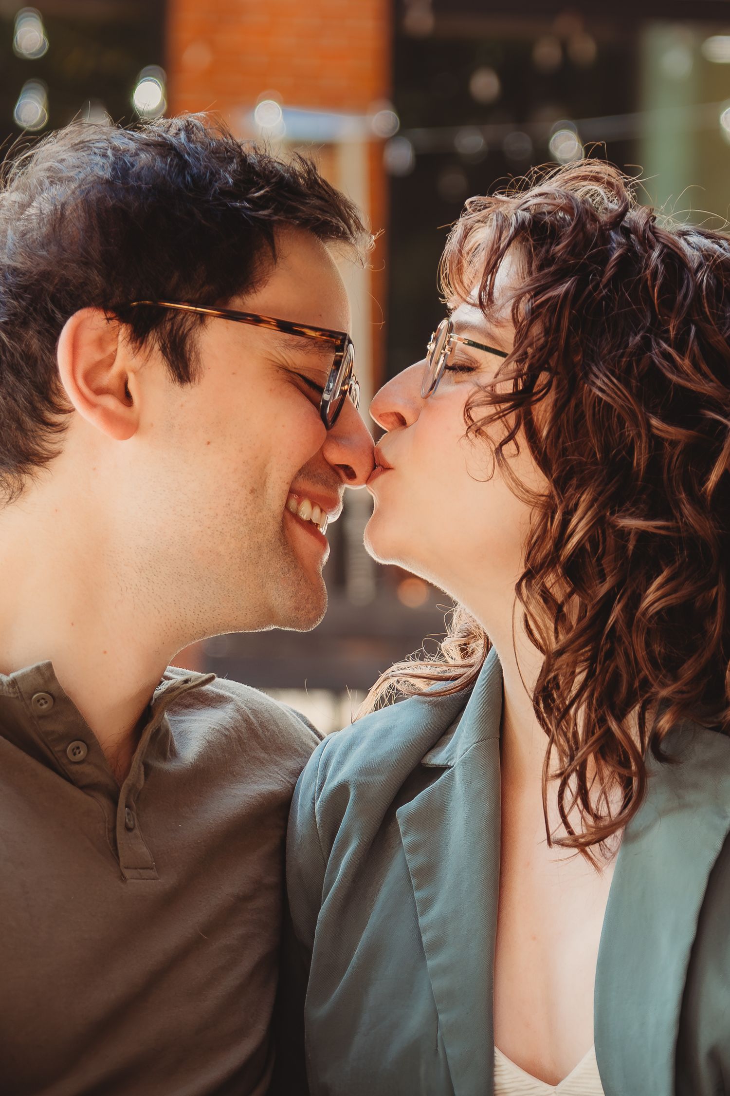 A smiling couple wearing glasses share a romantic kiss outdoors in warm sunlight.