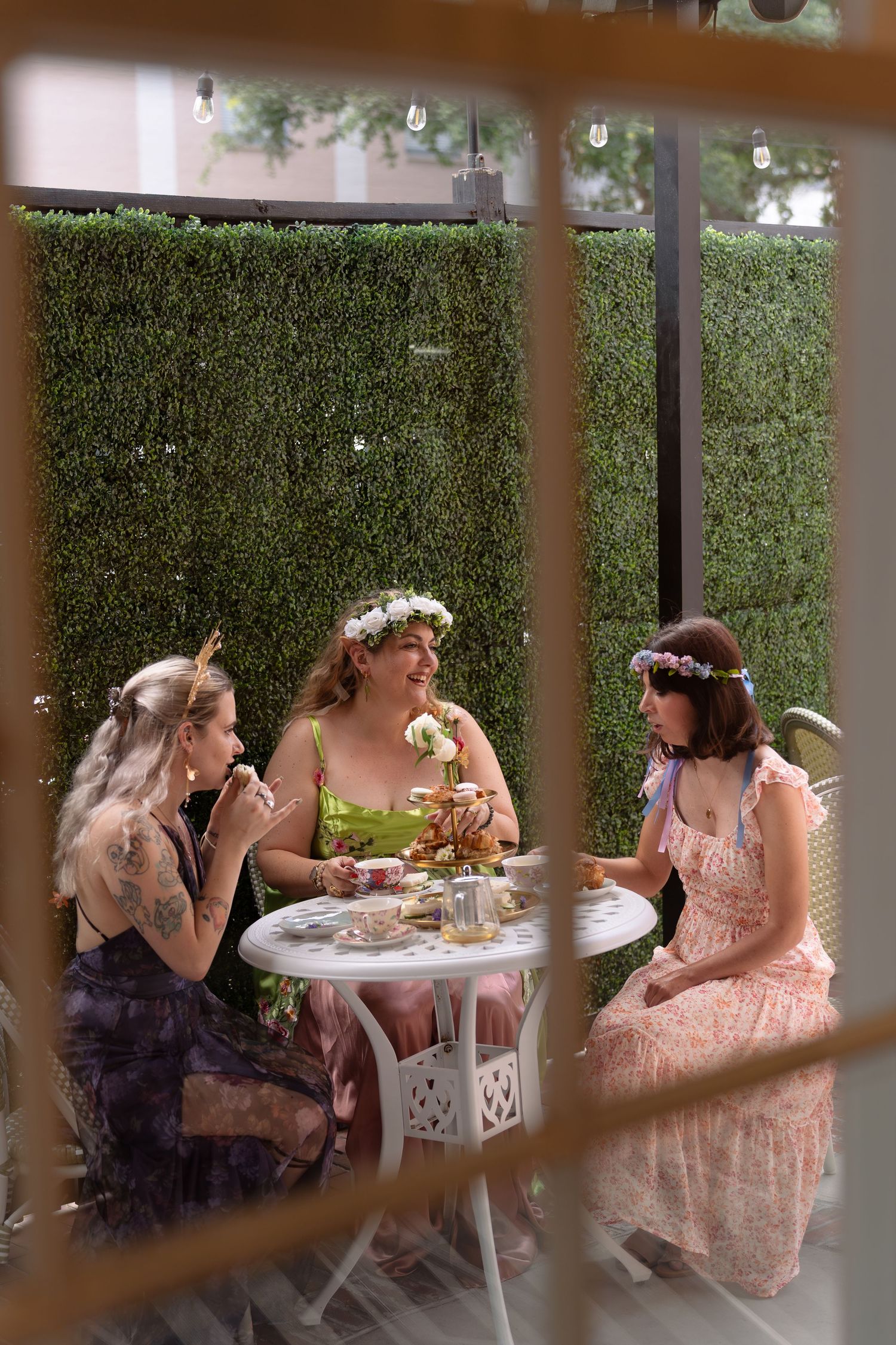 Three women wearing flower crowns enjoying tea party at outdoor garden table.