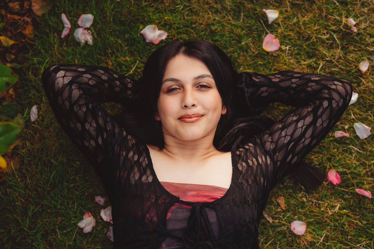 Young woman with dark hair lying on grass surrounded by scattered pink flower petals, smiling.