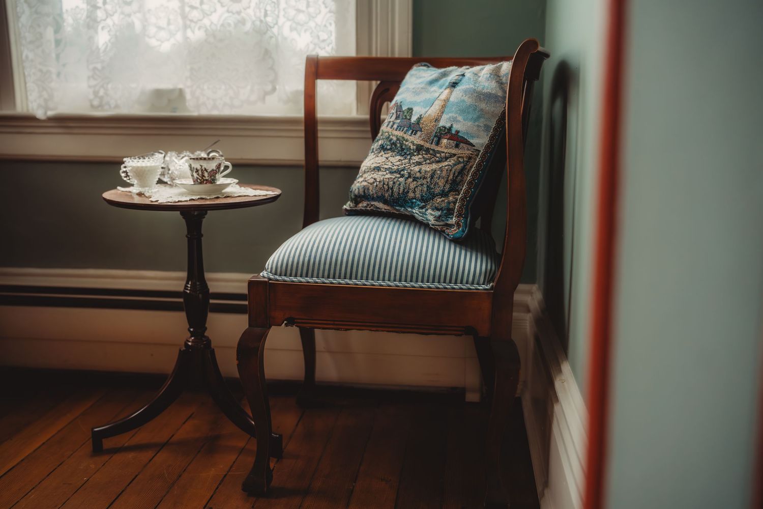Antique wooden chair with striped cushion and decorative pillow beside a small pedestal table with tea set near a window.