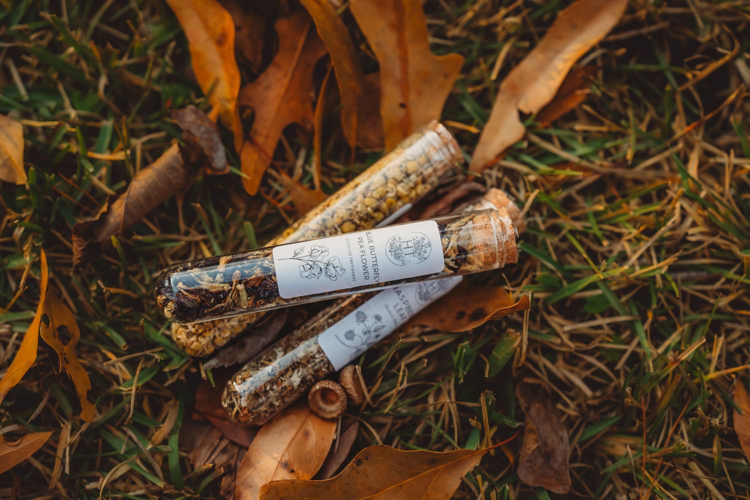 Three herbal smudge sticks with labels resting on autumn leaves and grass outdoors.