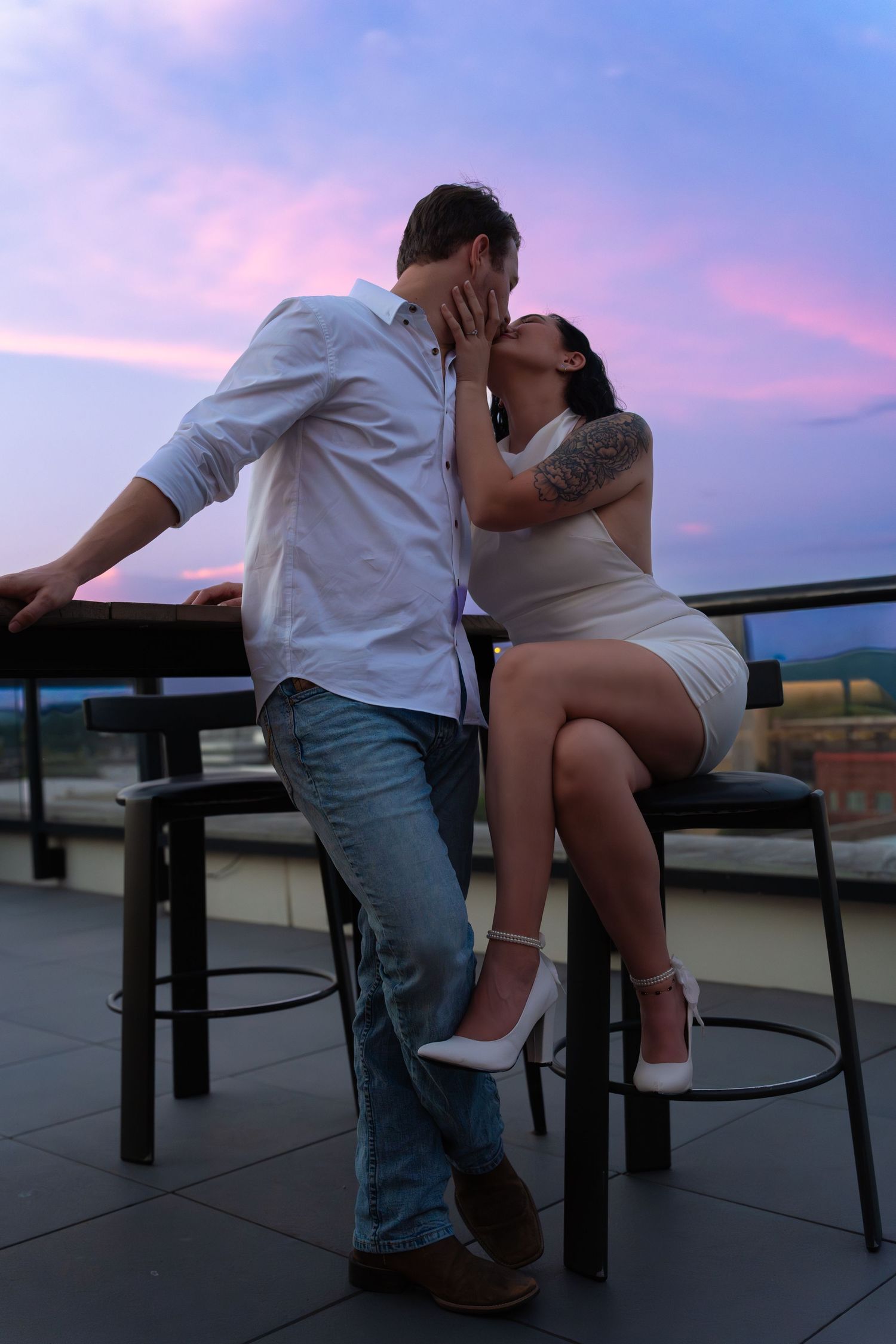 A couple shares a romantic kiss on a rooftop terrace at sunset, seated by bar stools under a pink and purple sky.