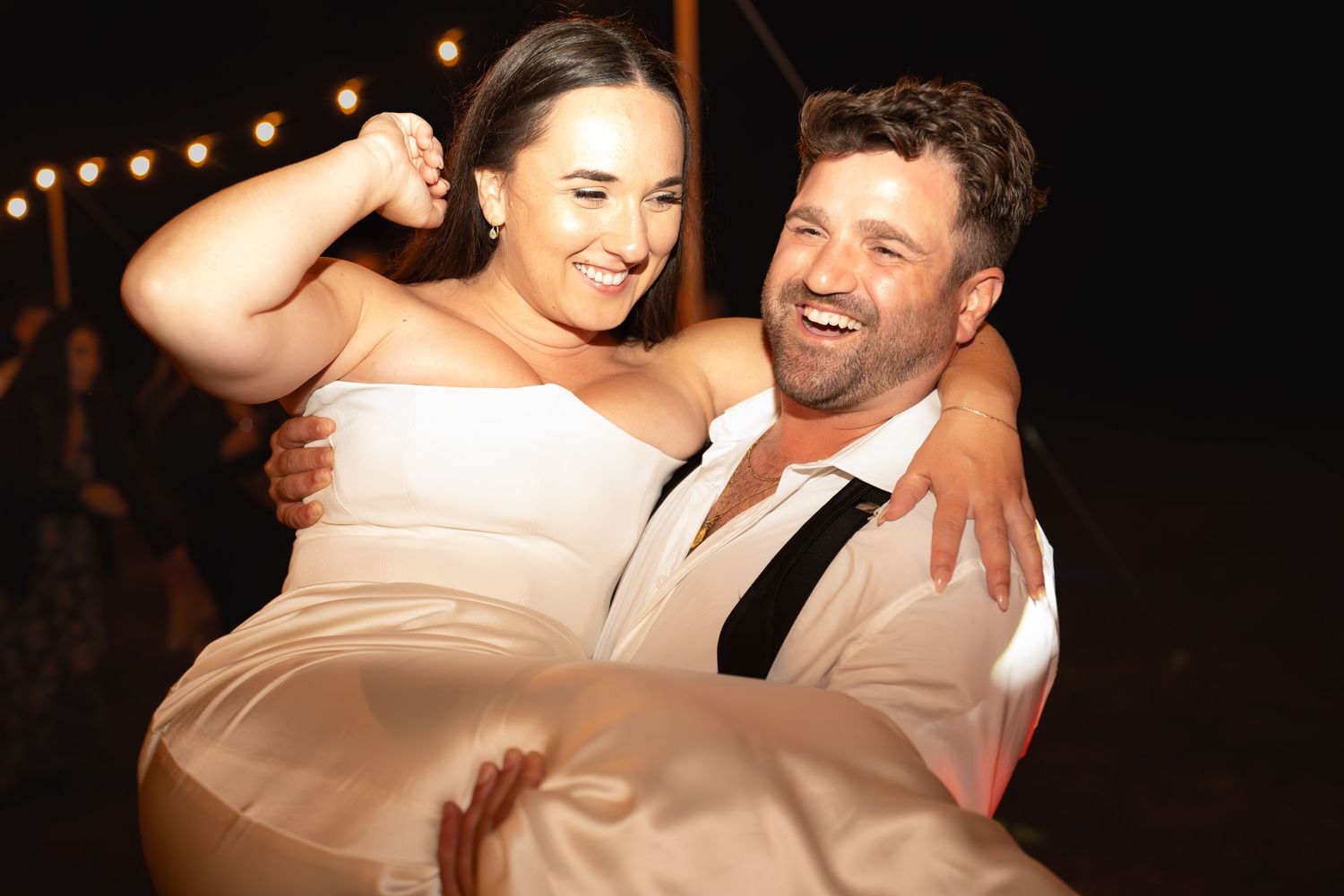 Groom carrying smiling bride in white strapless gown at outdoor wedding reception with string lights.