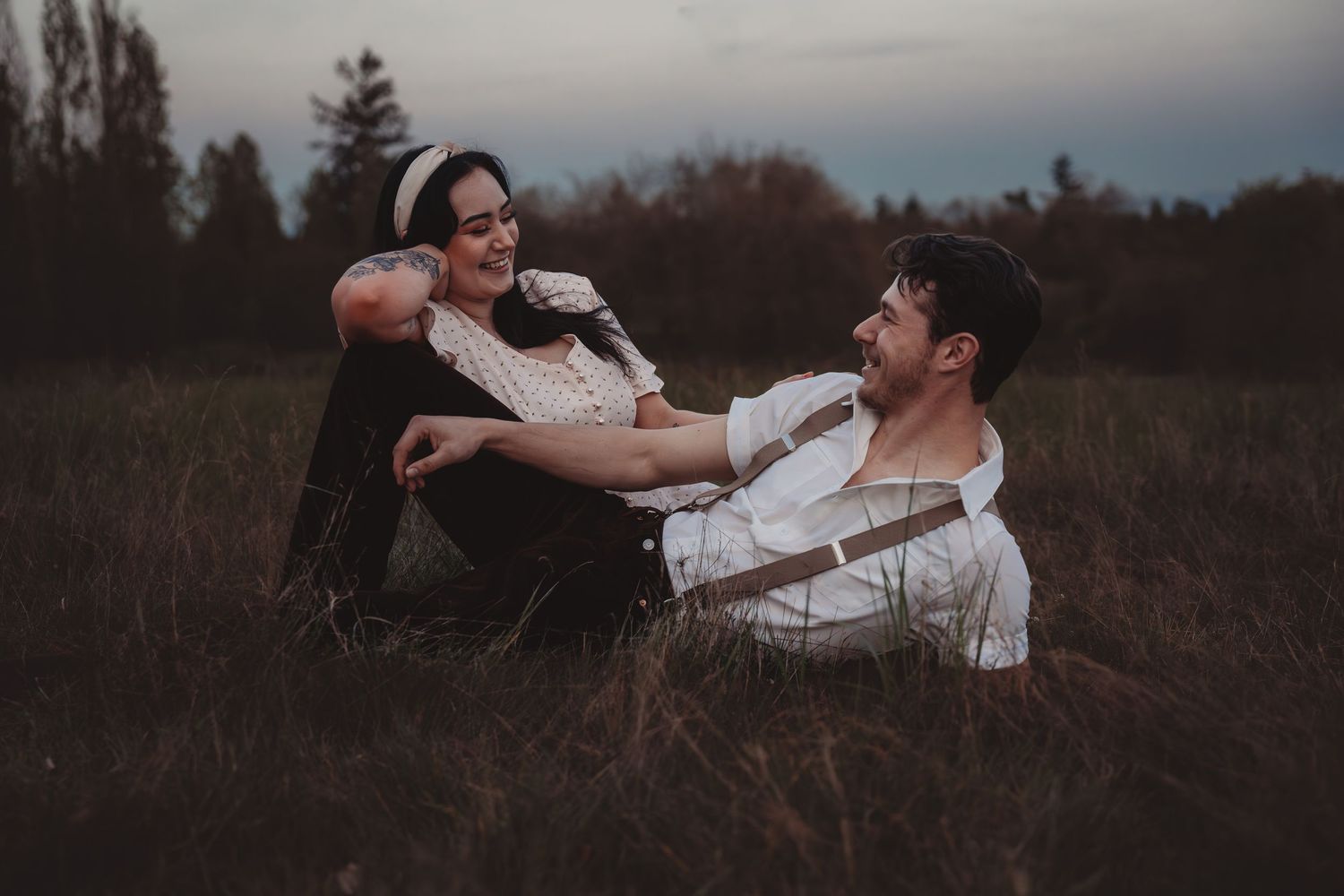 Couple sitting in a grassy field at dusk, smiling at each other in vintage-style white tops and dark pants.