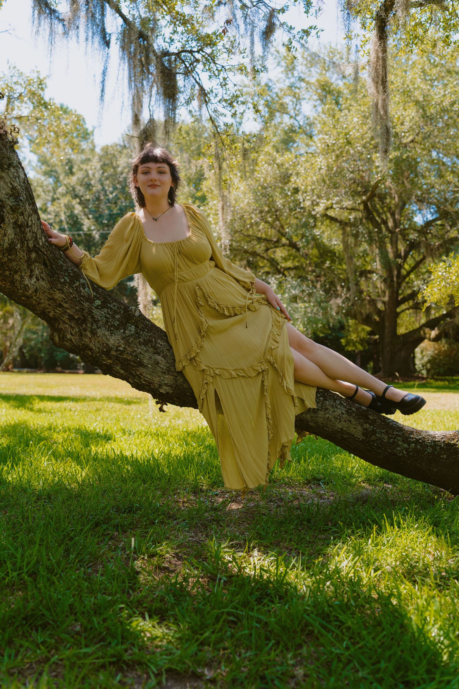 Woman in yellow dress sitting on tree branch in sunny park with Spanish moss-draped oak trees and green grass.