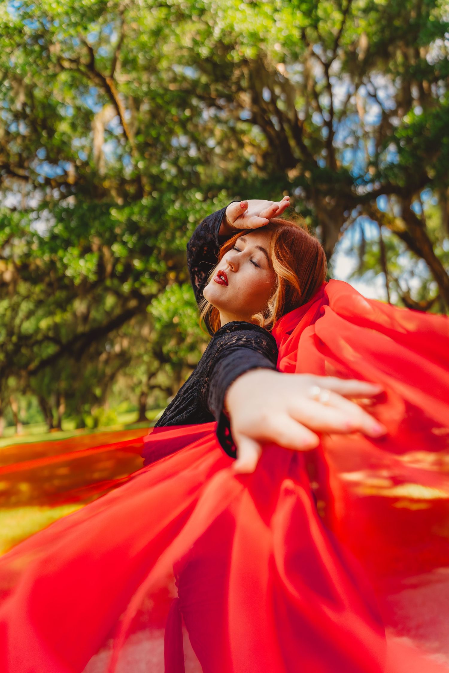 Woman in red flowing dress and black top poses dramatically outdoors among lush green trees.