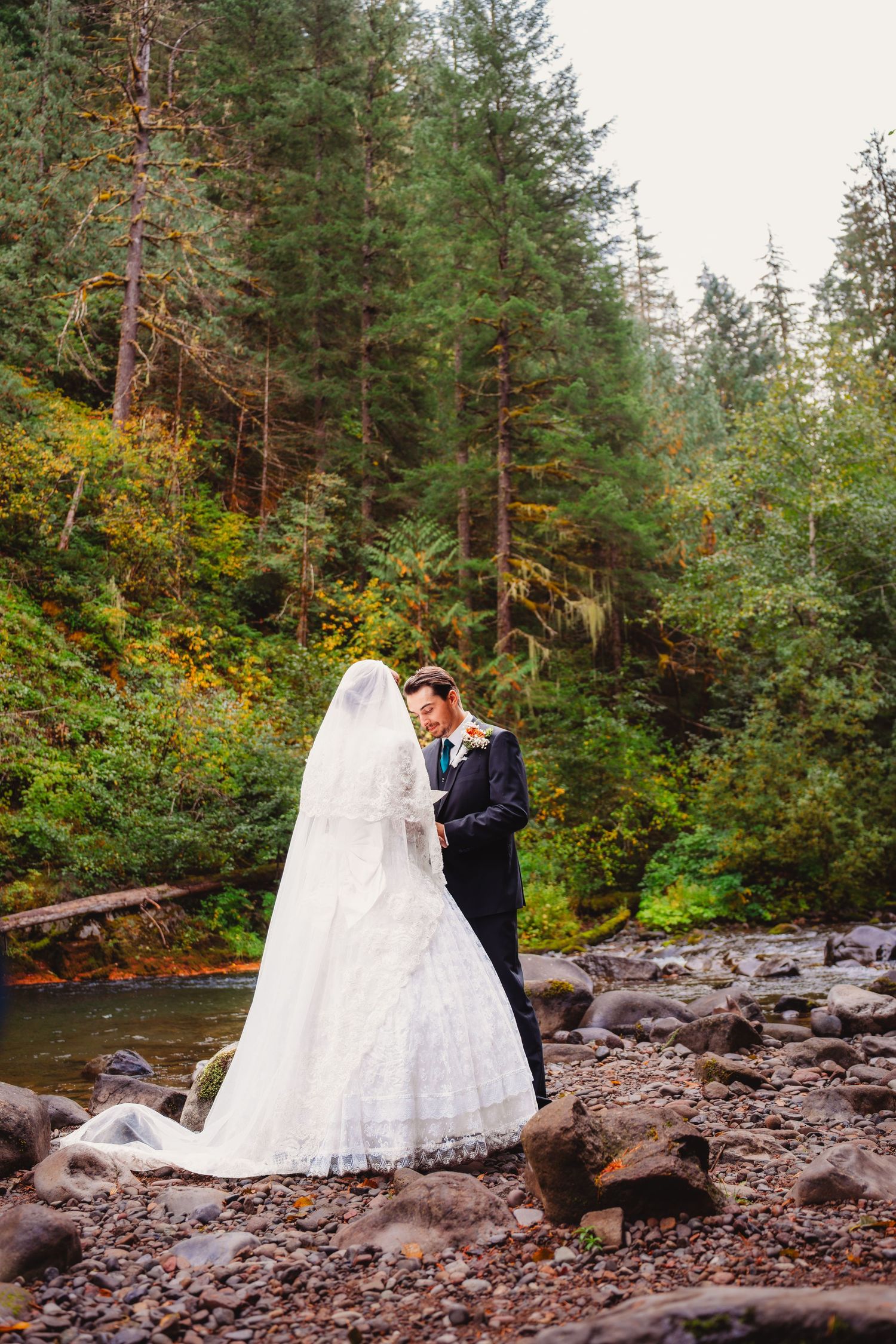 Bride and groom standing by a forest creek with tall pine trees and colorful autumn foliage. mt hood Oregon