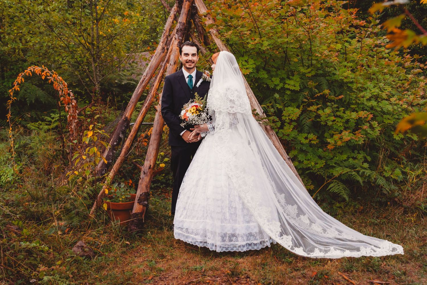 Bride and groom pose outdoors in autumn forest setting, bride wearing lace gown with long veil holding bouquet.