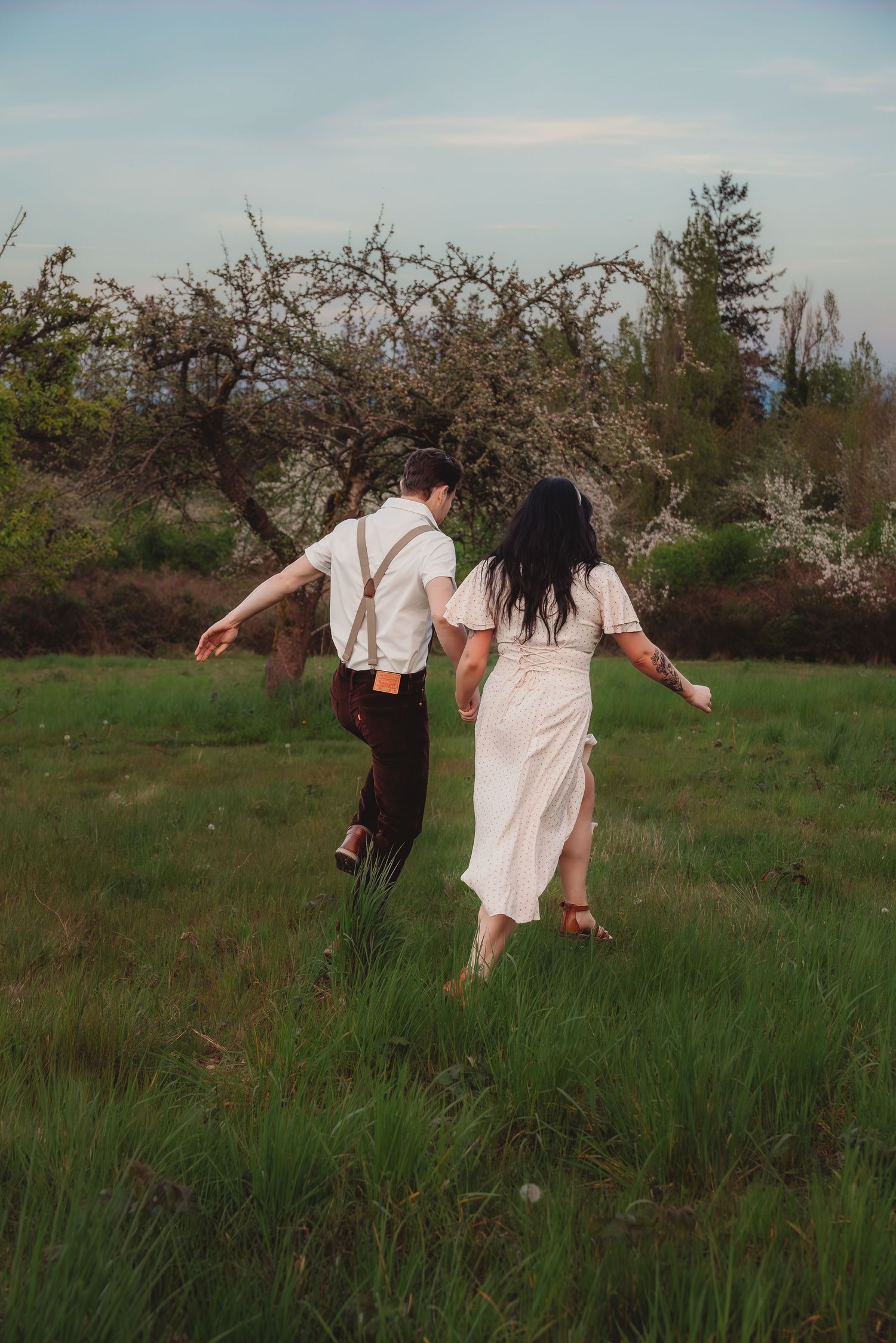 Couple holding hands running through green meadow, man in suspenders and woman in white dress, blooming trees background.