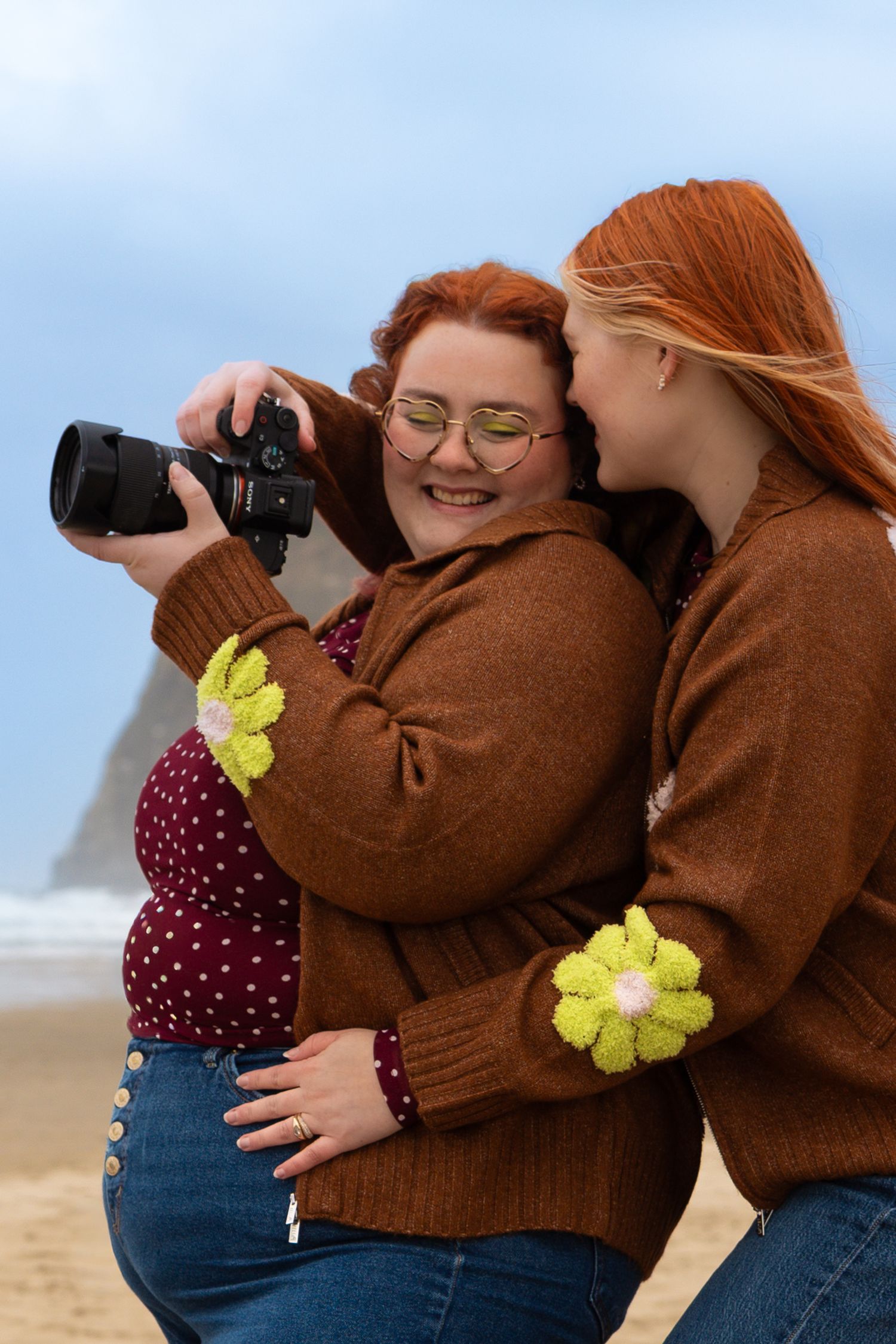 Two redhead women in matching brown sweaters with floral patches review photos on a camera at a beach.