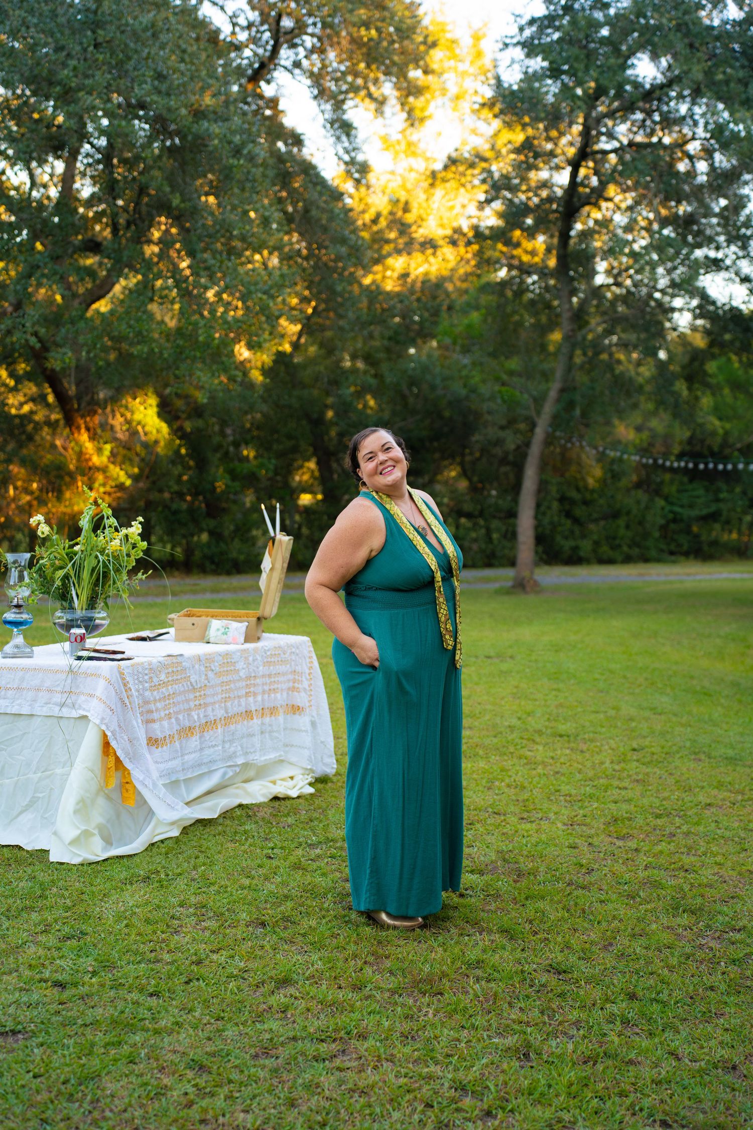 Smiling woman in a teal maxi dress stands on a grassy lawn near a decorated table with flowers, surrounded by trees at golden hour.