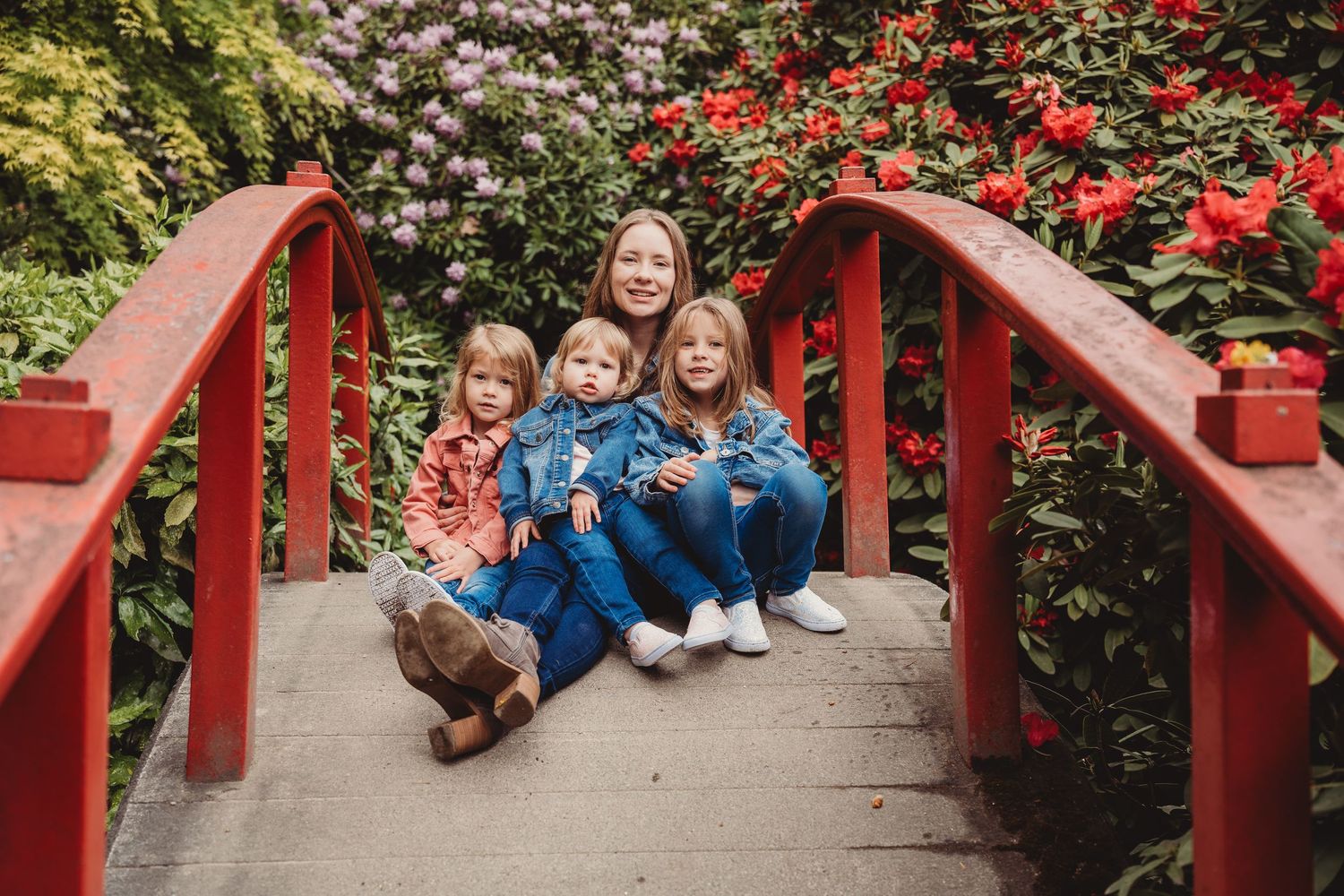 Four children sitting on a red bridge surrounded by colorful blooming flowers in a lush garden.