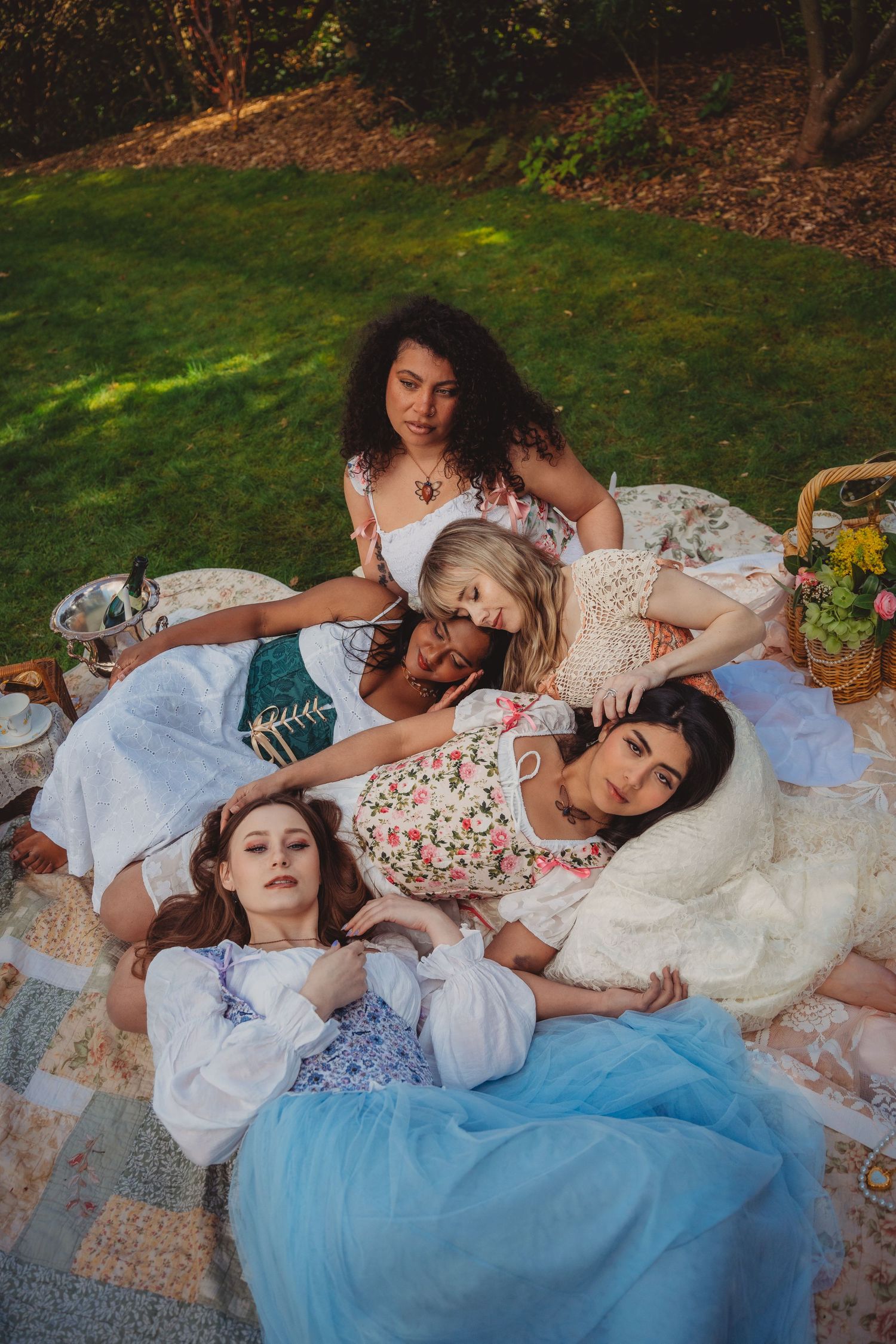 Five young women in floral and pastel dresses lounging together on a quilt blanket outdoors during a garden picnic.