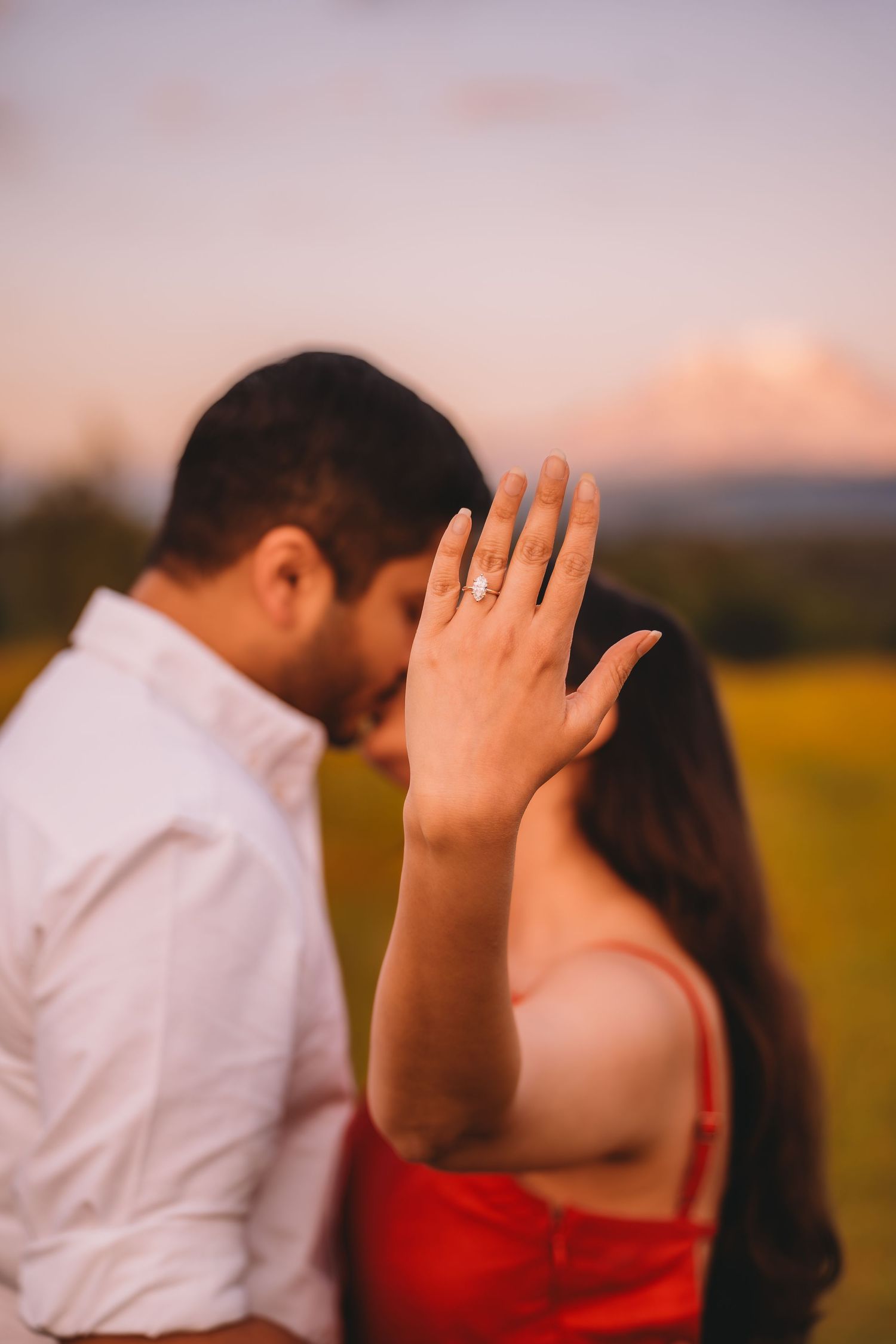 Couple sharing a romantic moment at sunset, woman showing off her engagement ring with a mountain in the background.