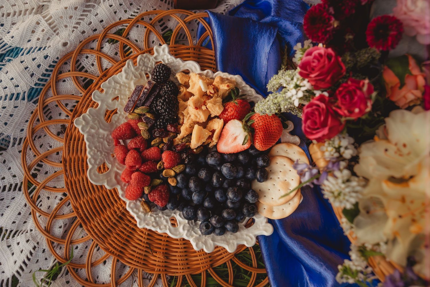 A wicker plate with fresh strawberries, blueberries, blackberries, and nuts beside colorful roses and flowers.