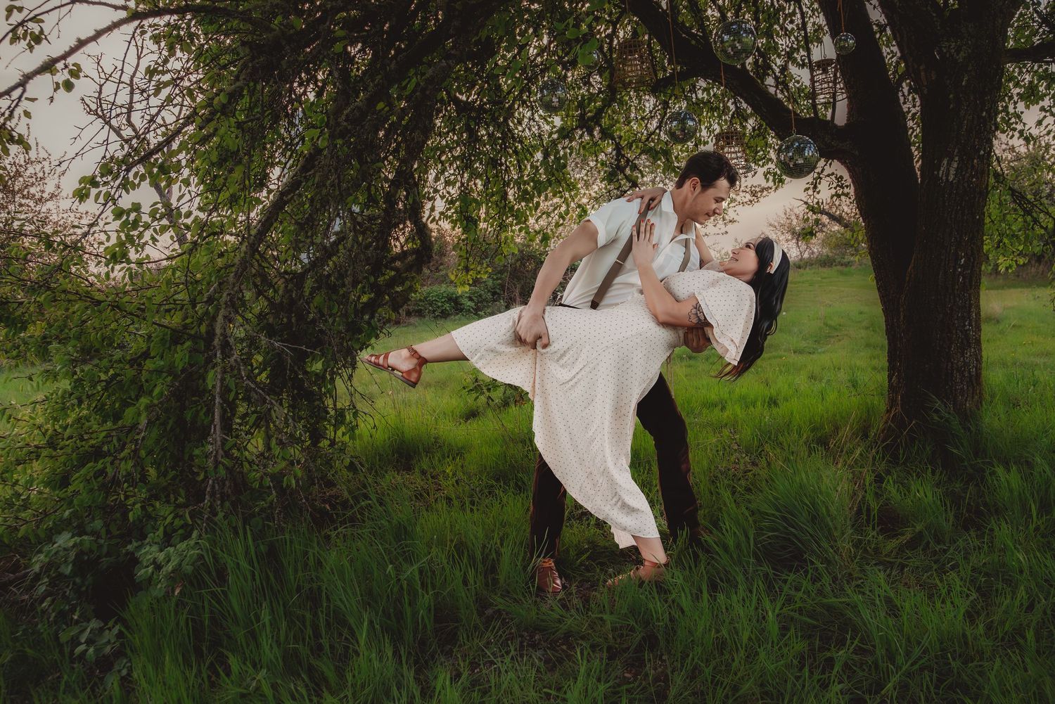 Groom lifting bride in white dress outdoors under green tree in lush garden during romantic wedding photo session.