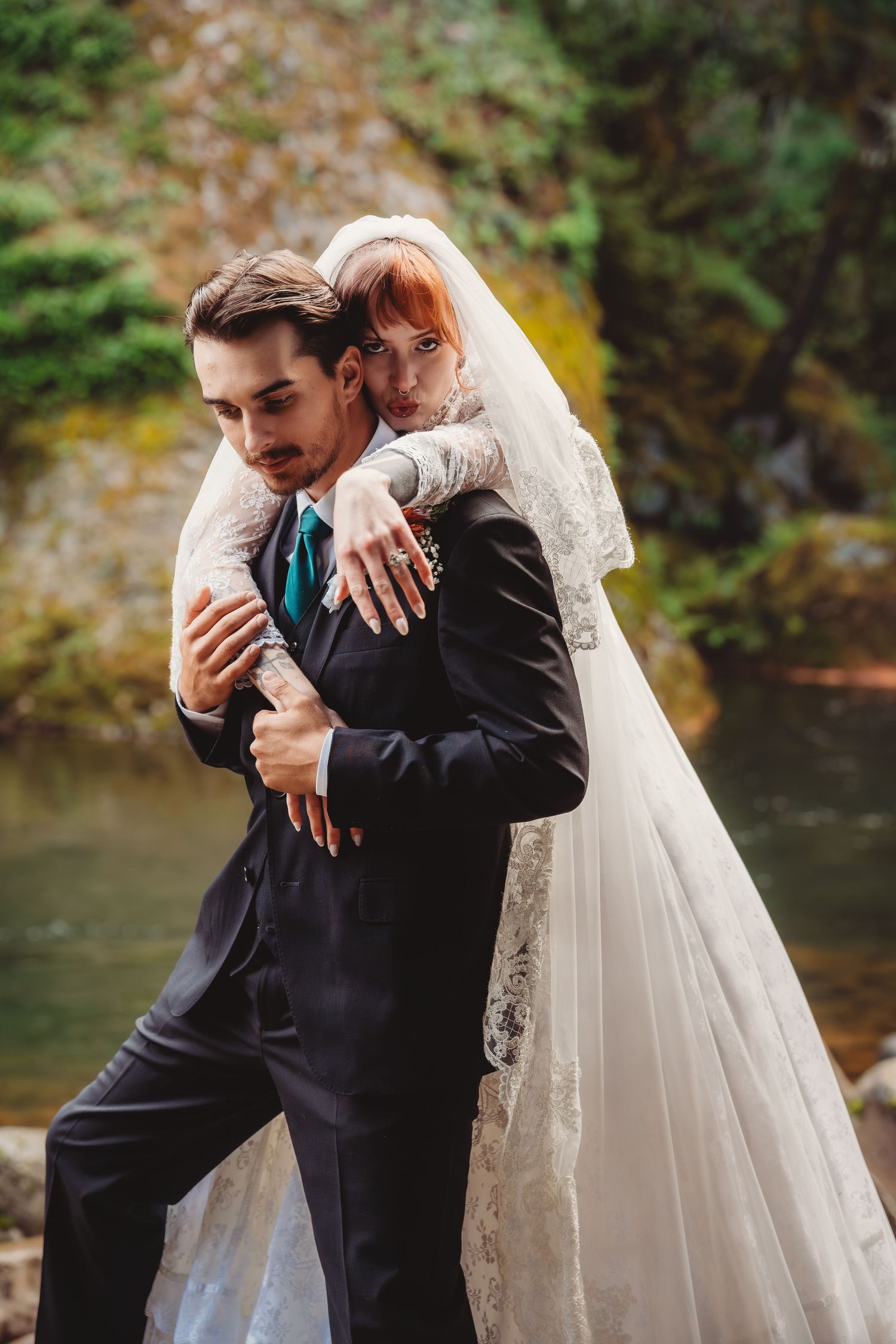 A bride in a lace gown and veil embraces her groom in a black suit outdoors near a forest stream.