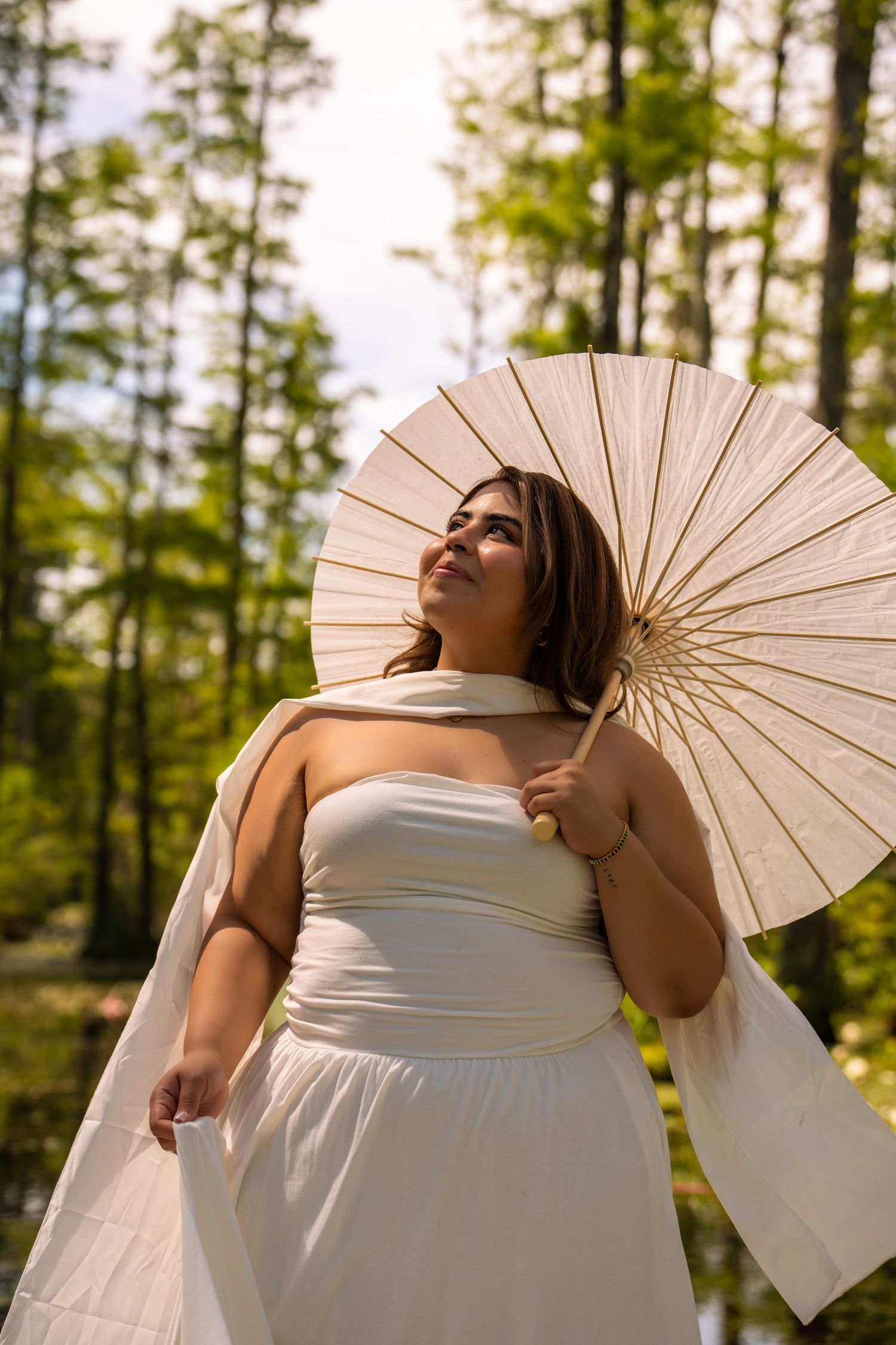 Woman in white dress holding a parasol umbrella outdoors in a lush green forest setting.