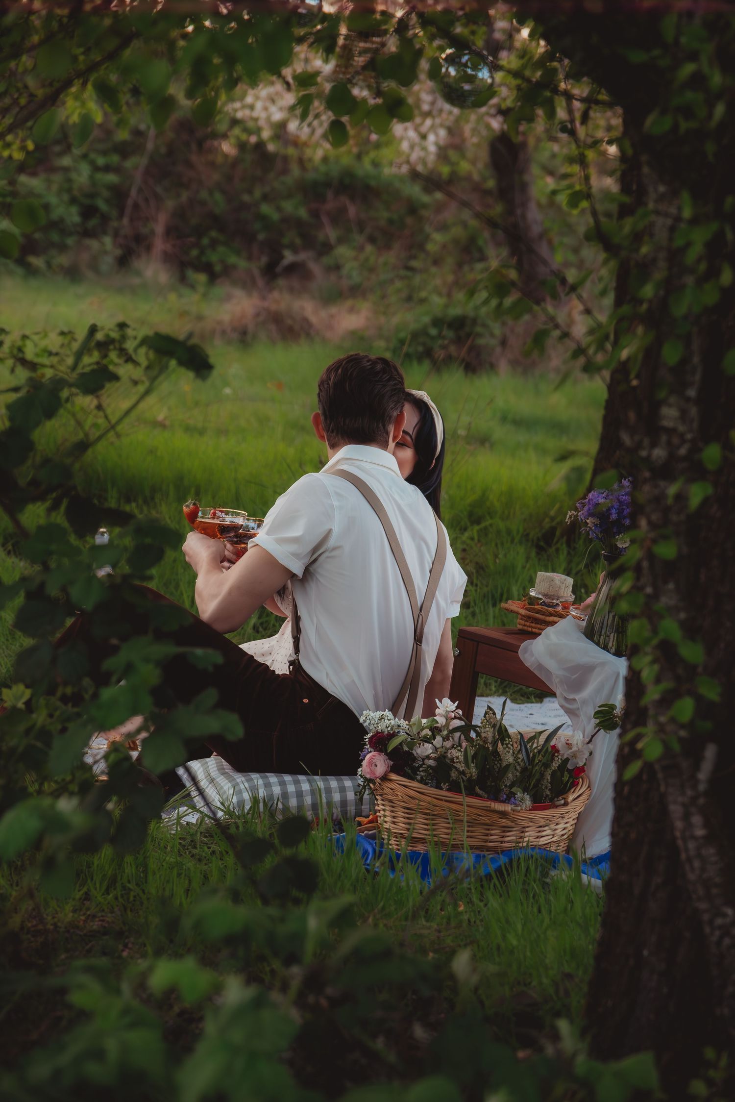 Couple enjoying a romantic outdoor picnic with wine and flowers in a lush green garden setting.