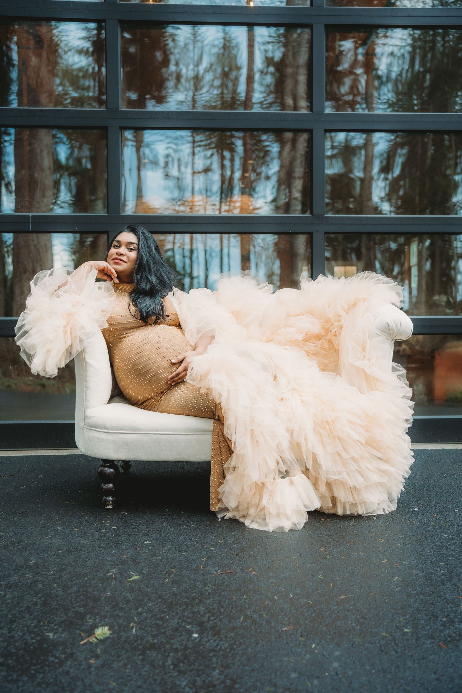 Pregnant woman in beige dress with peach tulle robe posing on white armchair.