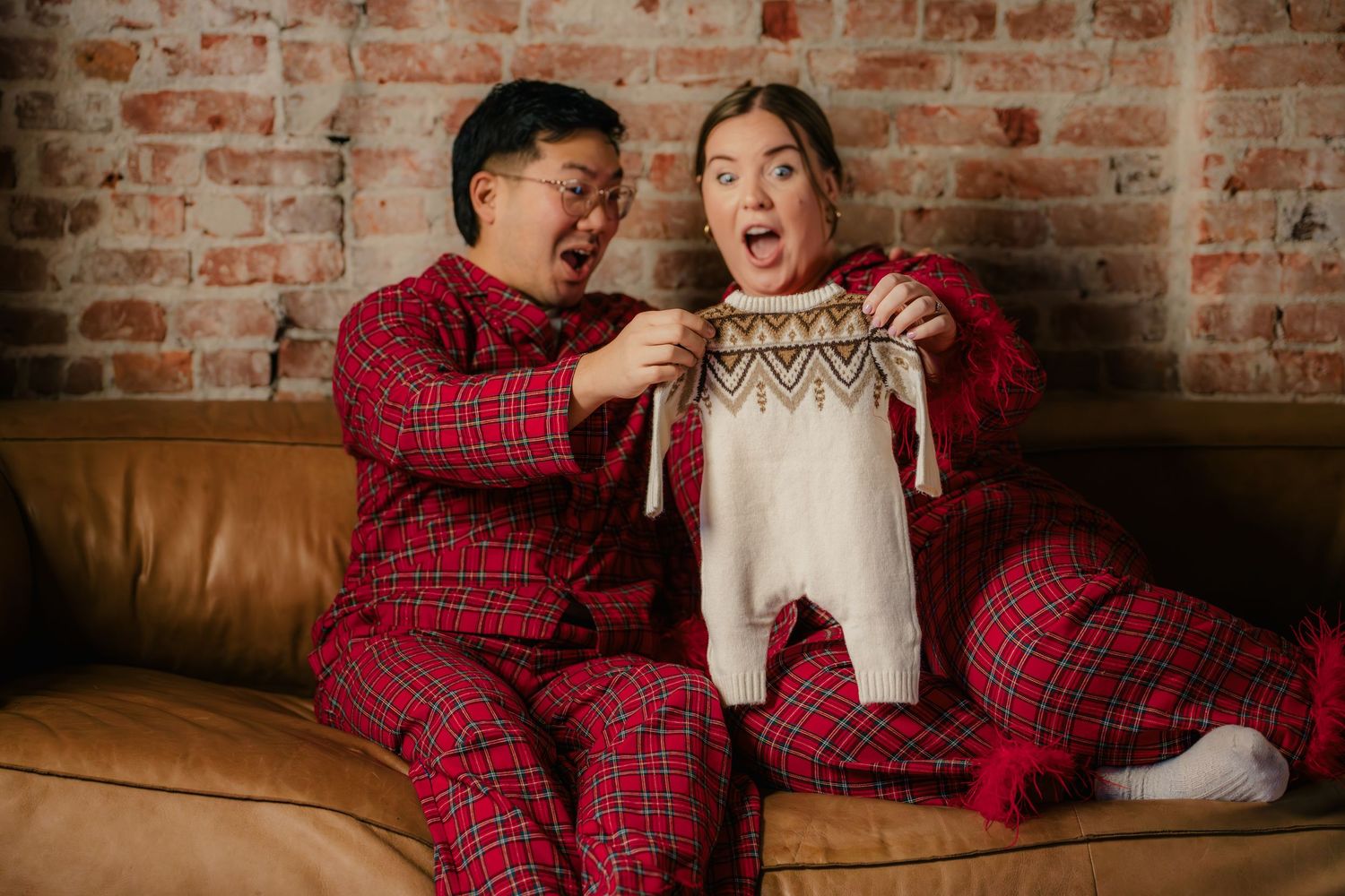 Couple in matching red plaid pajamas reacting with surprise to a baby onesie on a leather couch.