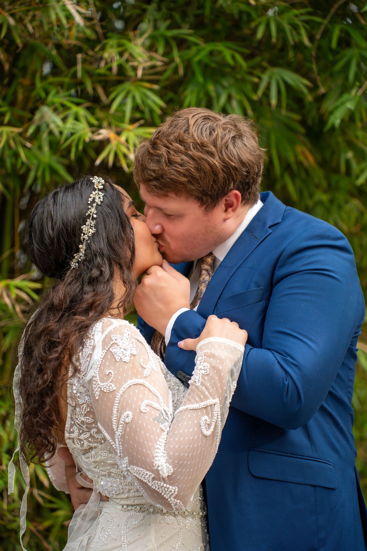 A bride and groom share their first kiss at the altar, surrounded by lush green bamboo foliage.