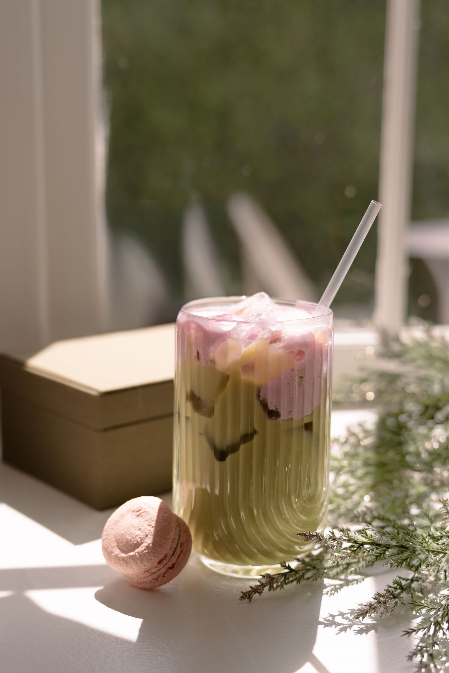 Iced matcha latte with pink cream, clear straw, pink macaron, and greenery on a sunlit windowsill.