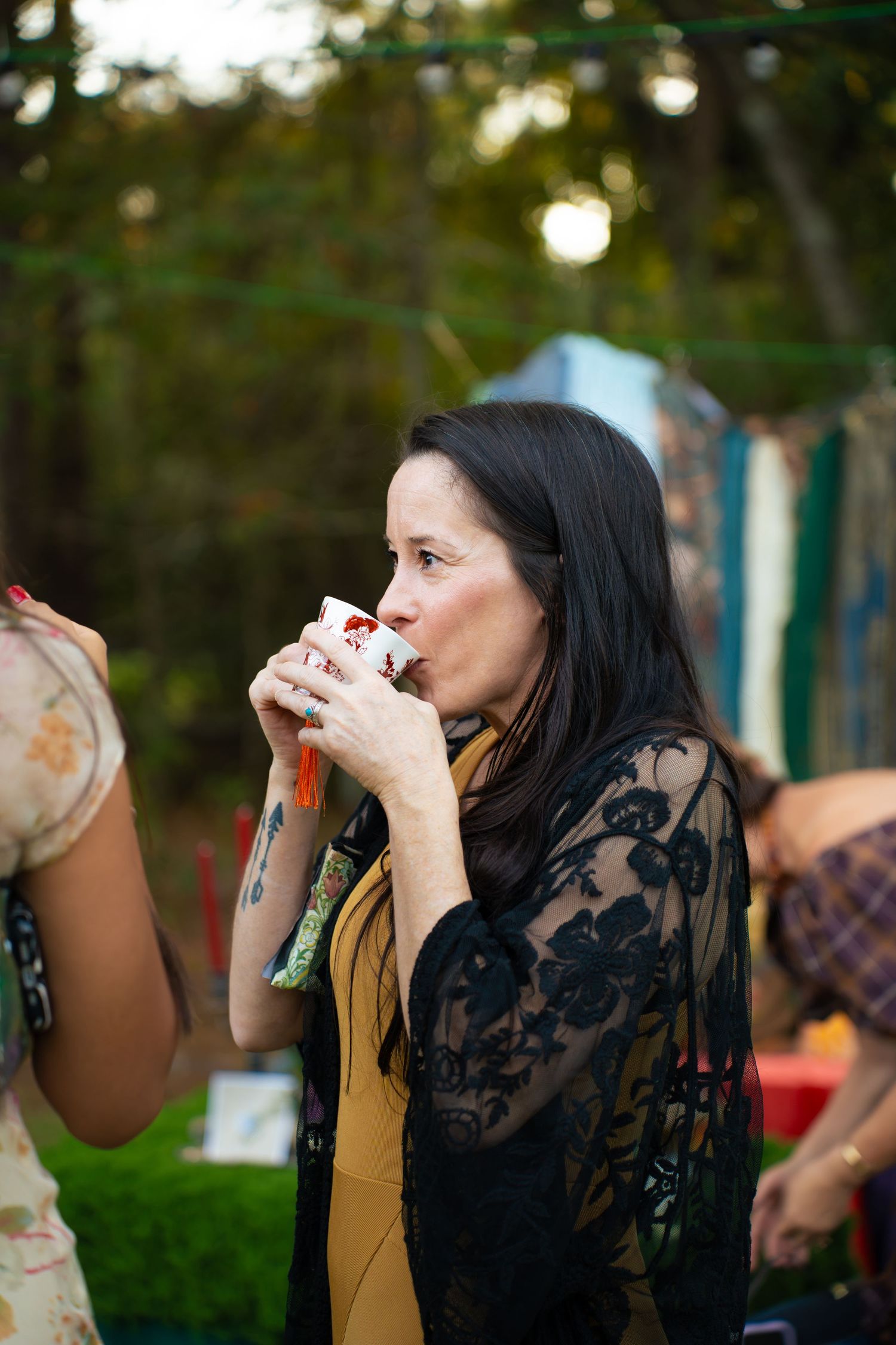 Woman with long dark hair and a black lace cardigan sips from a floral cup at an outdoor gathering among trees.