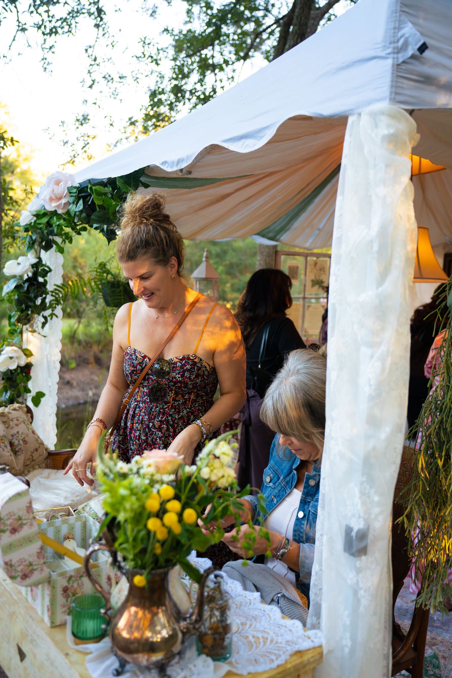 Smiling woman in a floral dress stands beside a table of yellow flowers in a brass pitcher under a white garden tent at sunset.