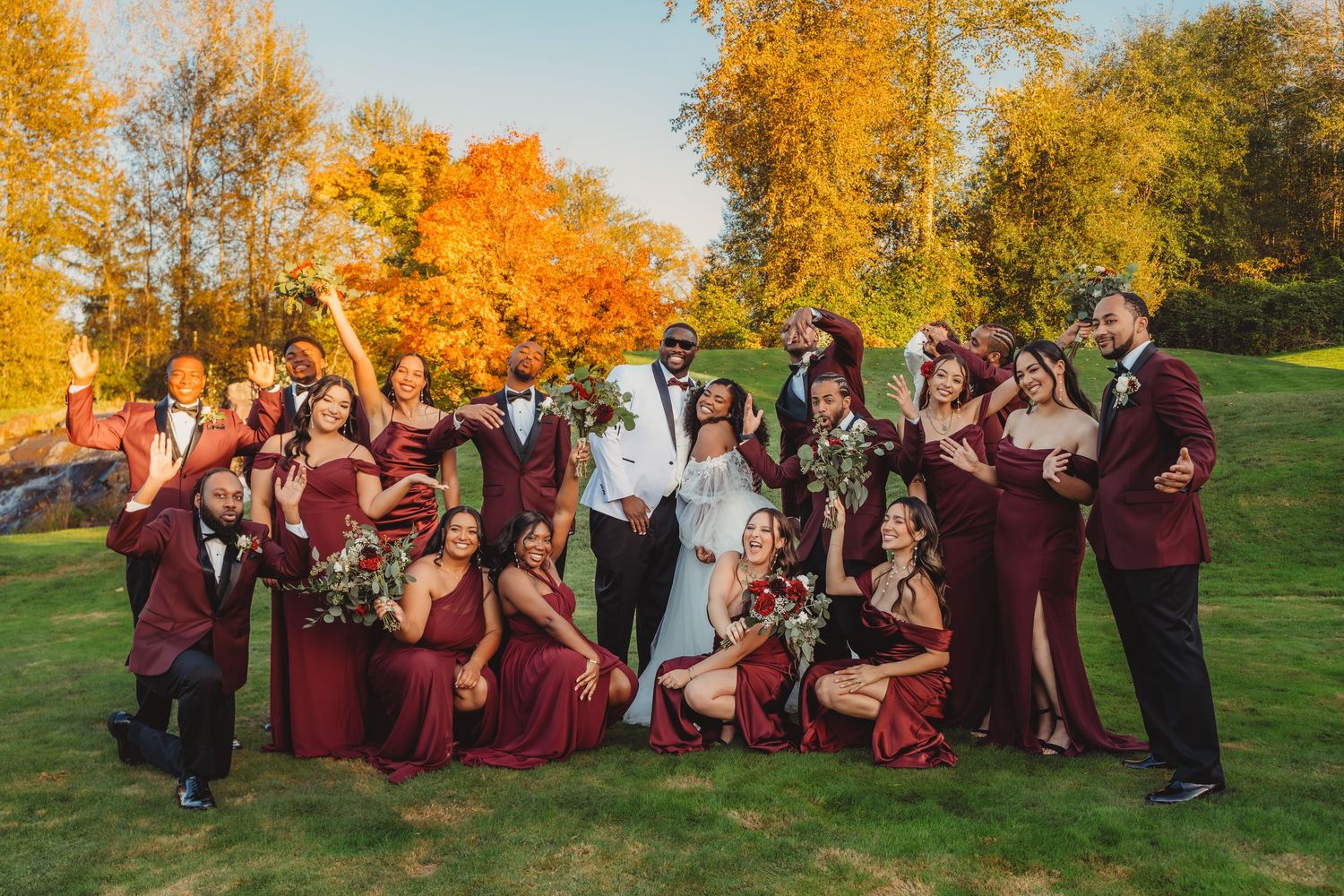 Wedding party in burgundy and white attire celebrating outdoors amid vibrant autumn foliage on a sunny day.