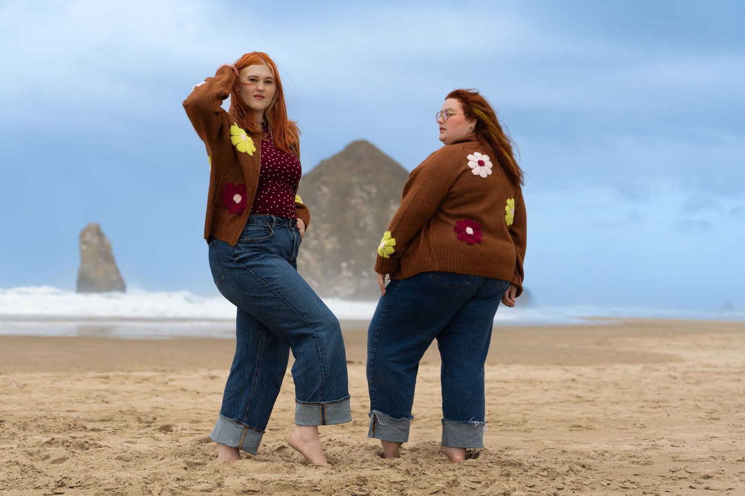 Two women in matching brown floral cardigans and jeans pose on a sandy beach near Haystack Rock.