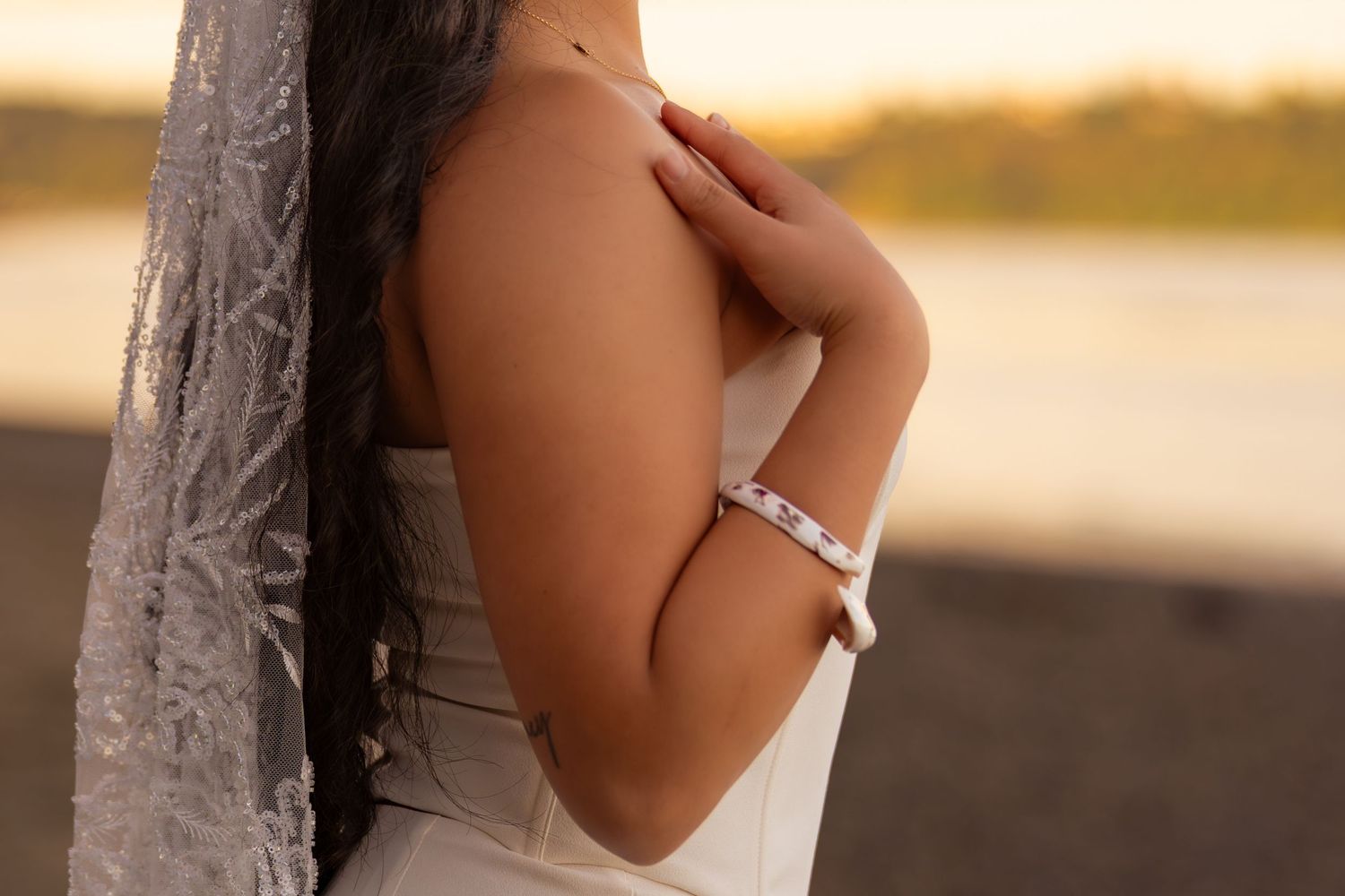 Bride in white dress and veil at sunset beach, showcasing elegant diamond bracelet and bare shoulder.