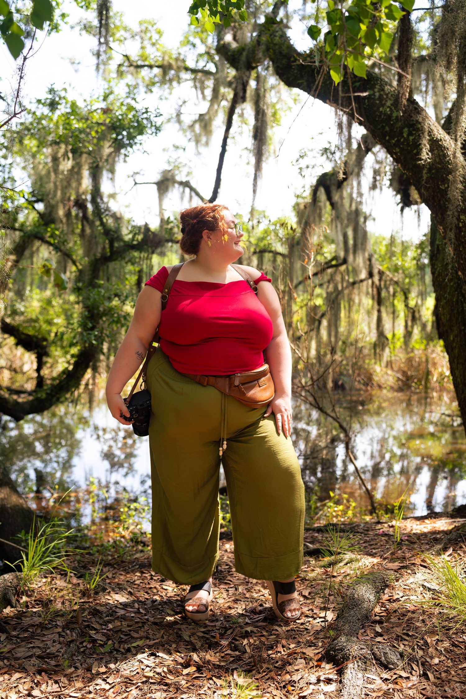 Plus-size woman in red top and olive pants standing under Spanish moss-draped trees near a tranquil pond.