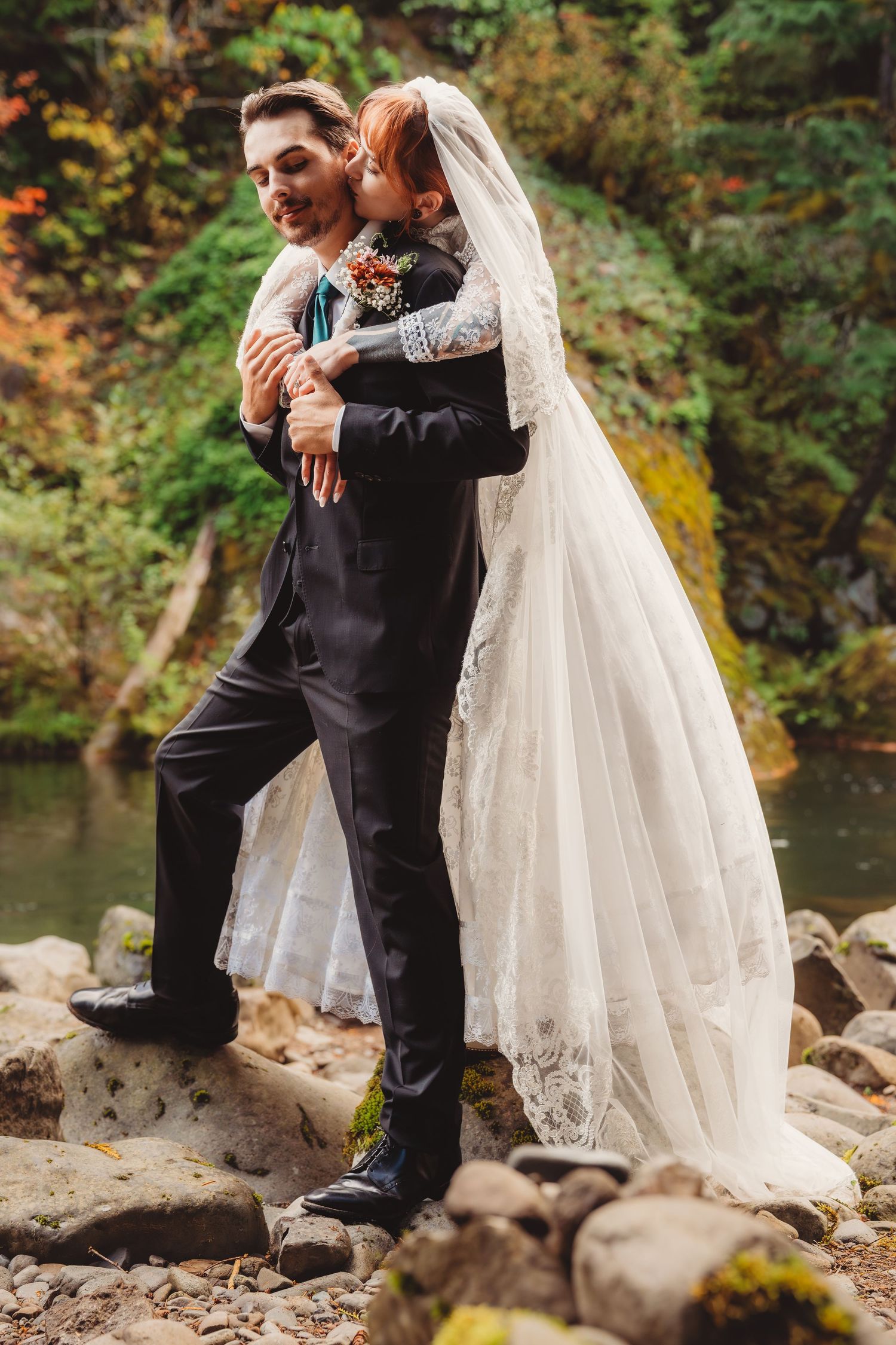Bride in lace wedding dress and veil embraces groom in black suit on rocky riverbank surrounded by autumn foliage.