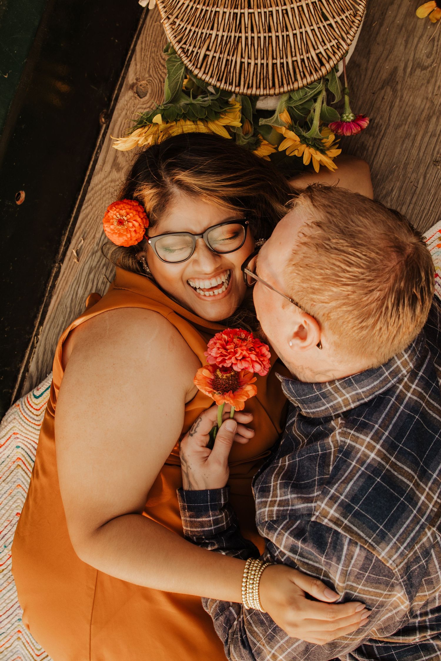 Couple laughing and lying down together, woman wearing glasses holding orange zinnia flowers in a warm autumn-themed setting.