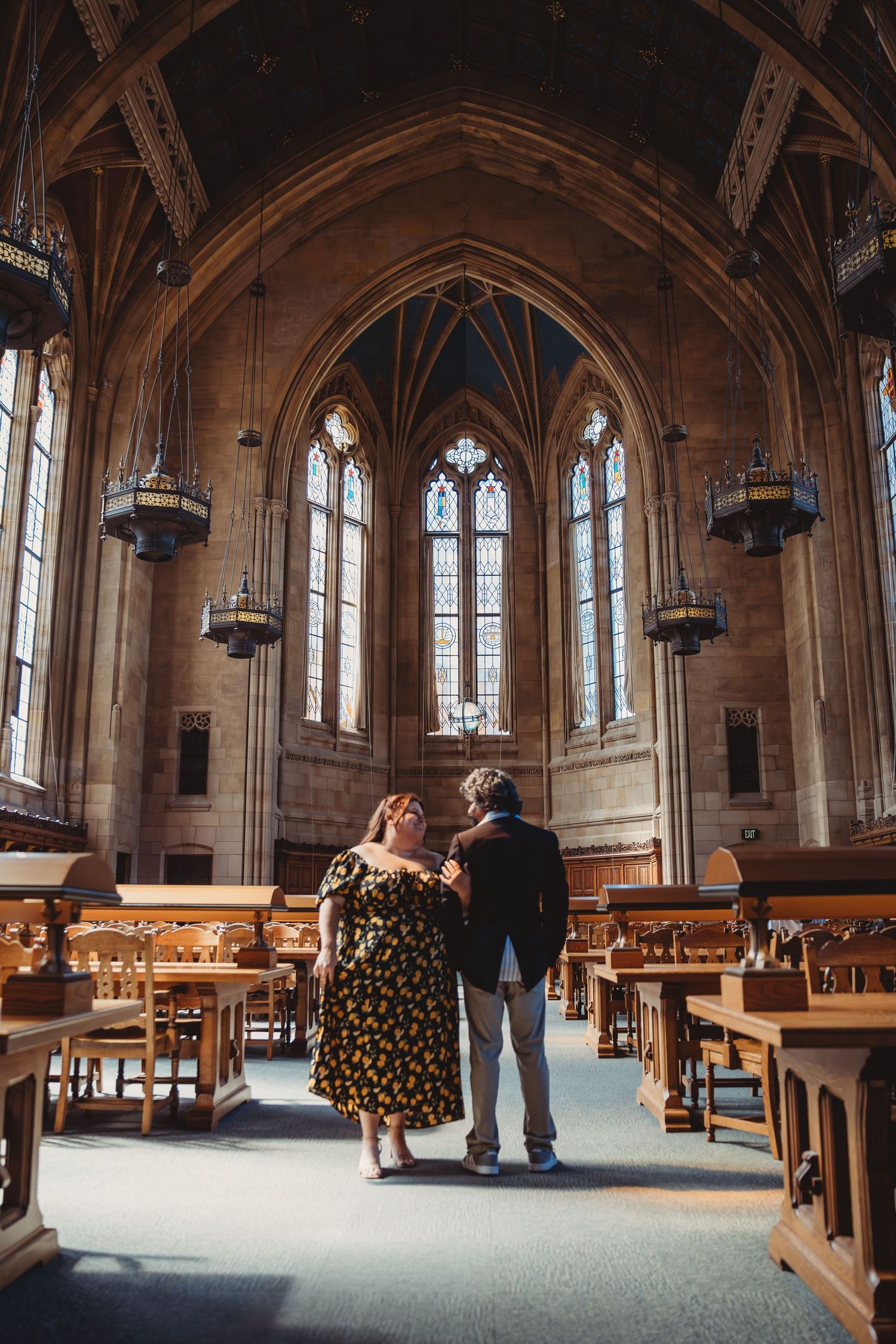 Couple embracing in a stunning Gothic cathedral library with arched ceilings, stained glass windows, and wooden pews.