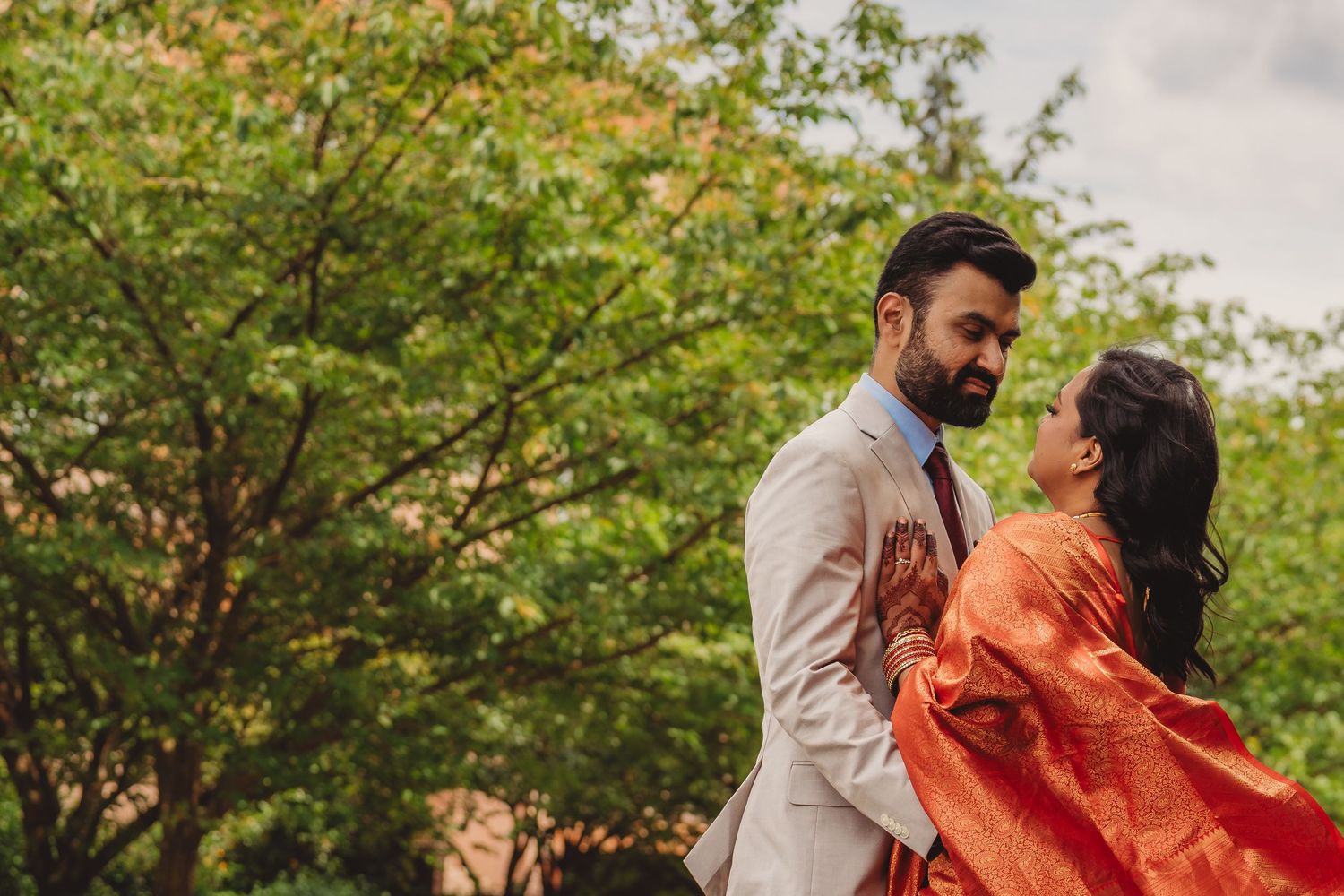 Couple in elegant attire embracing outdoors, man in white suit and woman in orange saree, with lush green trees behind.