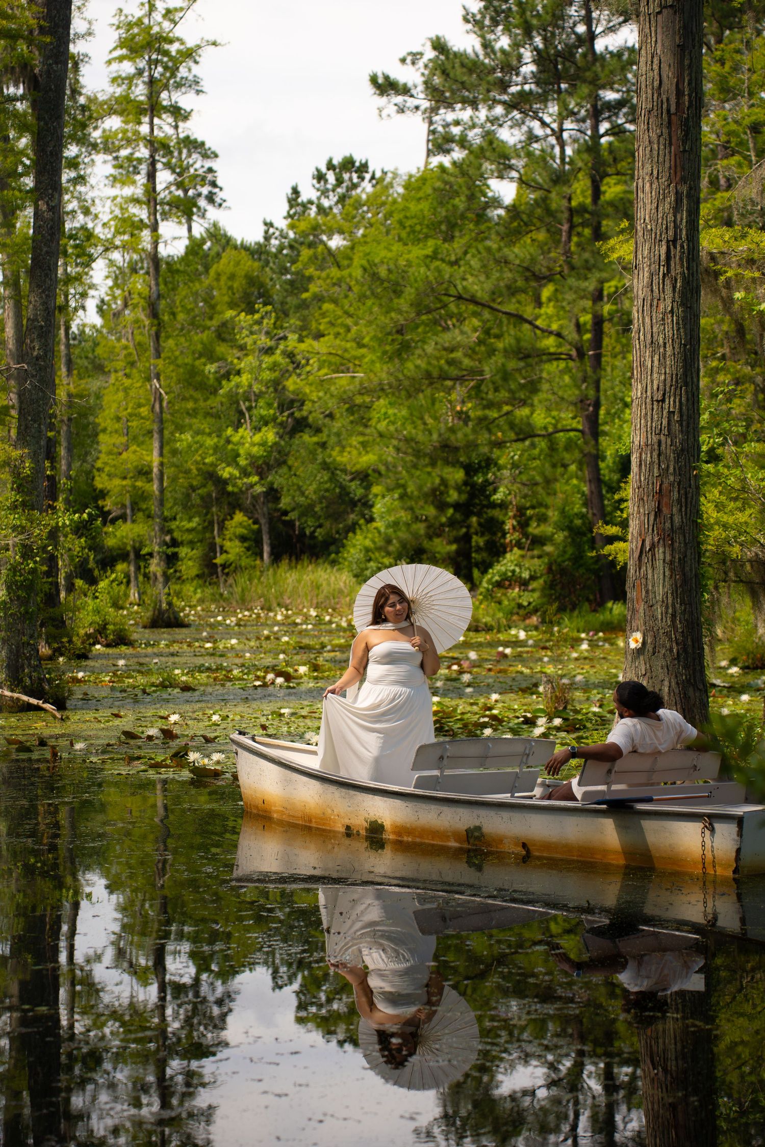 Bride in white gown holding parasol sits in rowboat on serene lily pad lake surrounded by lush green trees.